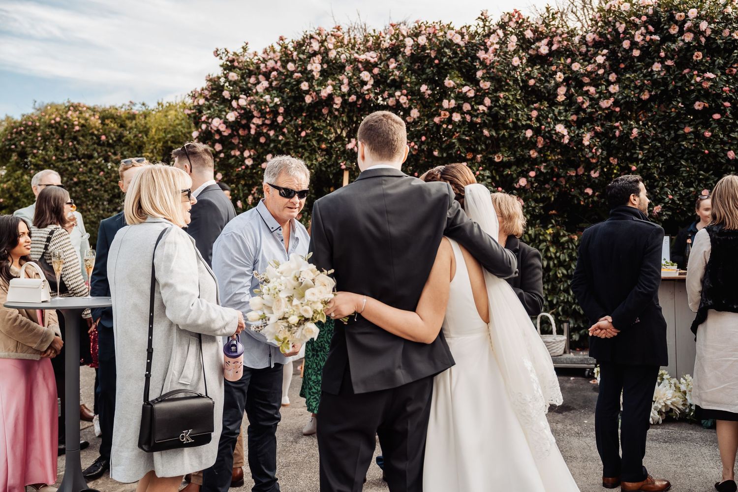 Wedding guests mingling outdoors near blooming floral bushes during a garden reception.