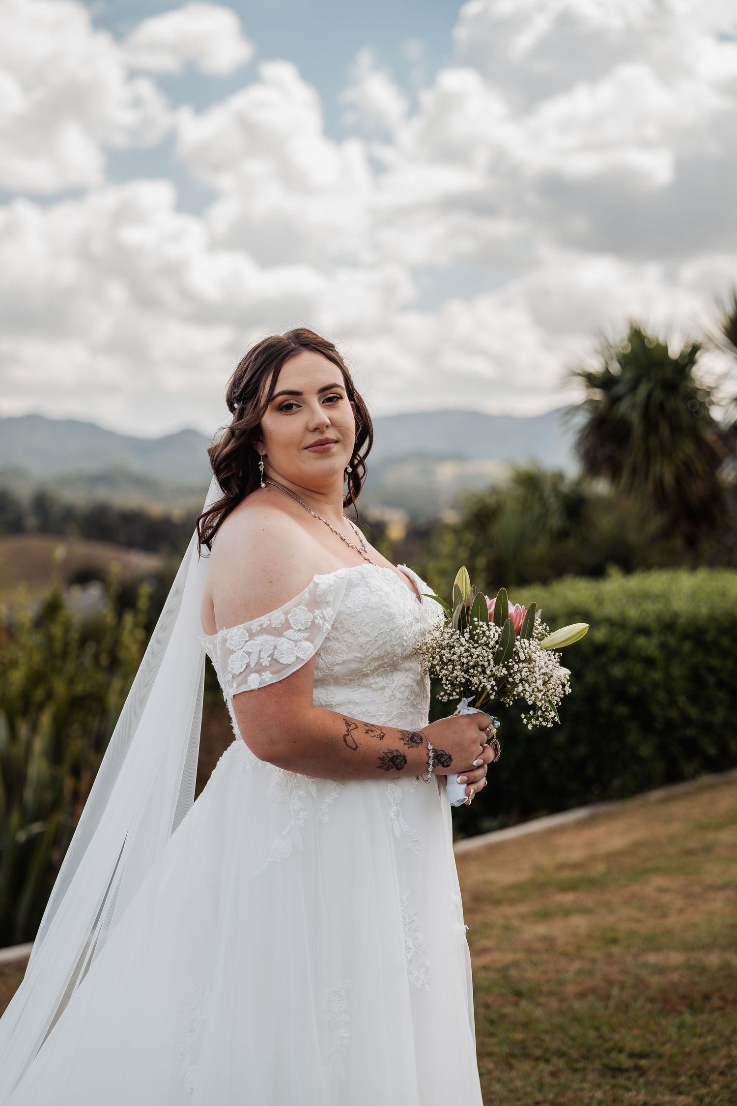 Elegant bride in off-shoulder lace wedding gown with veil holds bouquet outdoors with mountain views.