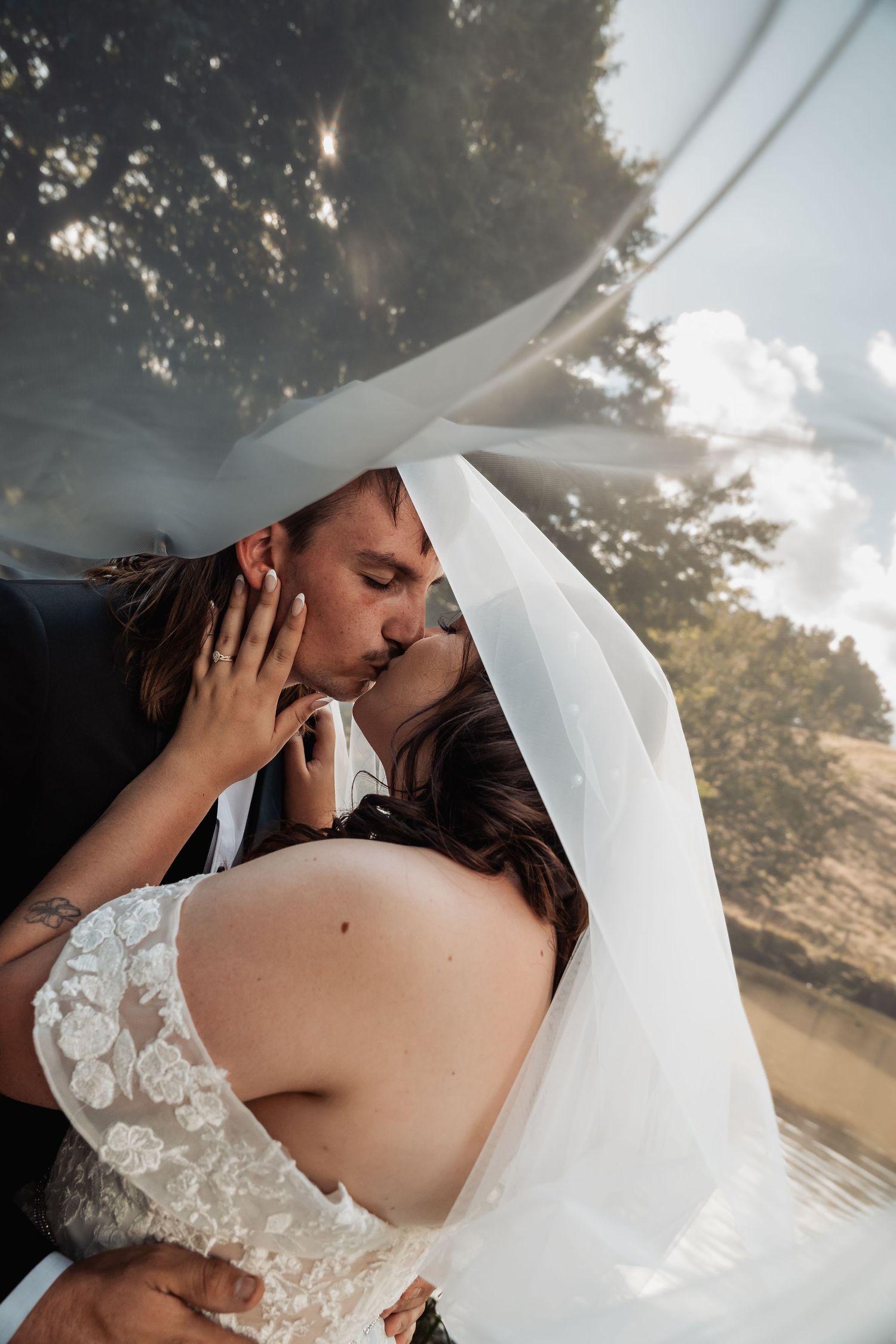Bride and groom sharing romantic kiss under flowing white veil with sunlit trees in background.