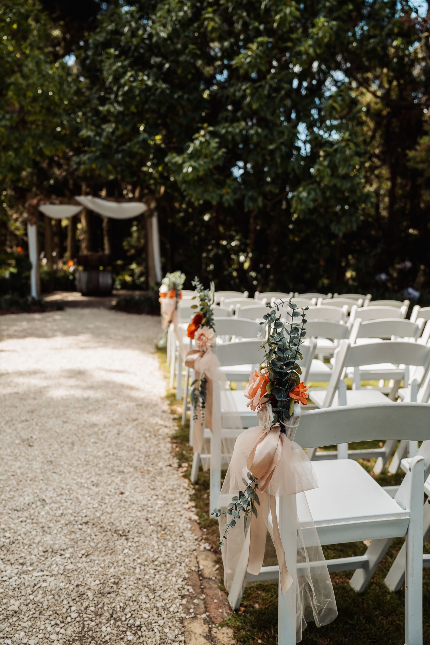 White folding chairs line a gravel aisle decorated with orange floral bouquets and blush ribbon at an outdoor wedding ceremony.