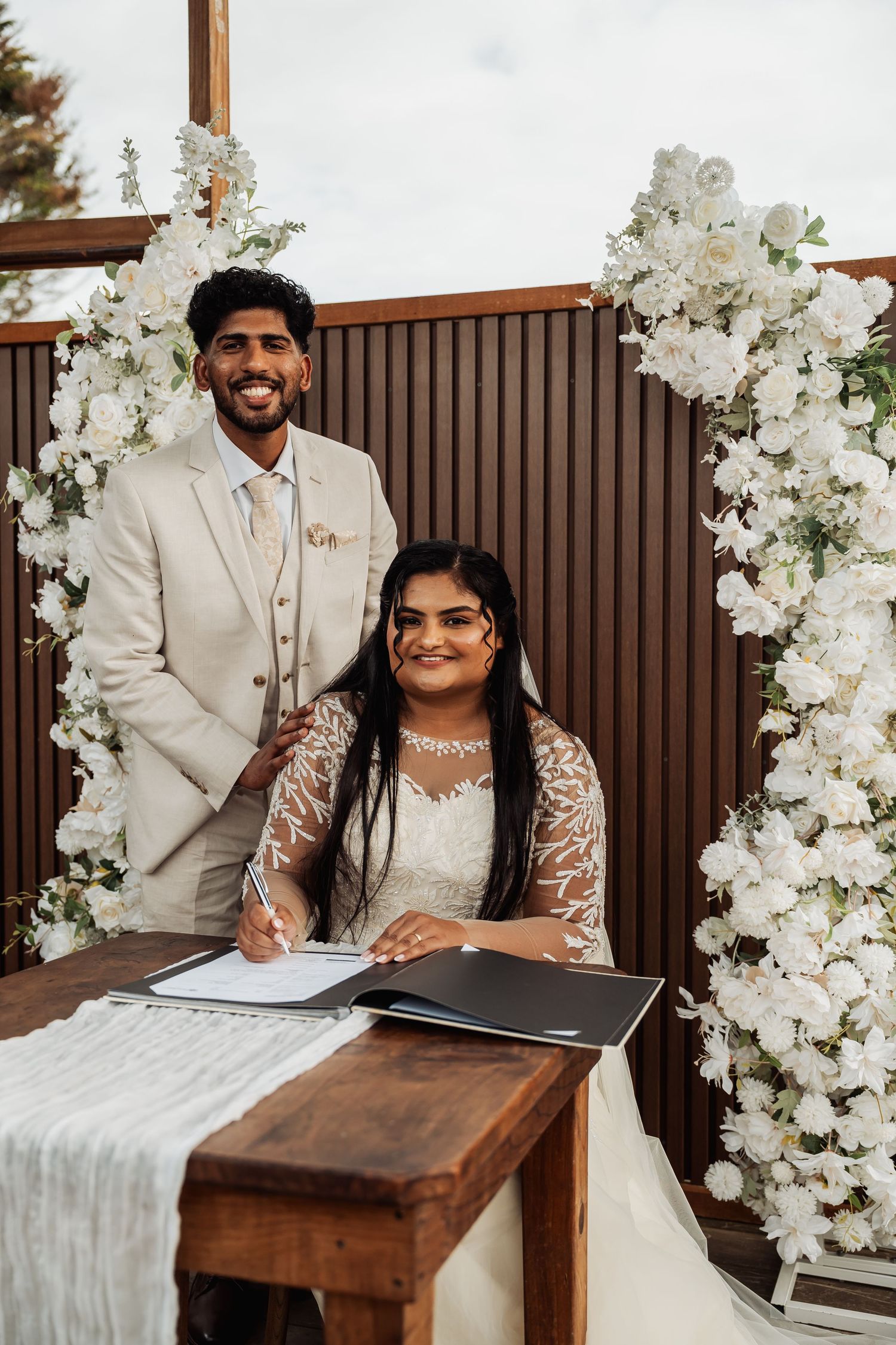 Bride signs wedding register at rustic outdoor ceremony with white floral arch while groom stands smiling behind her.