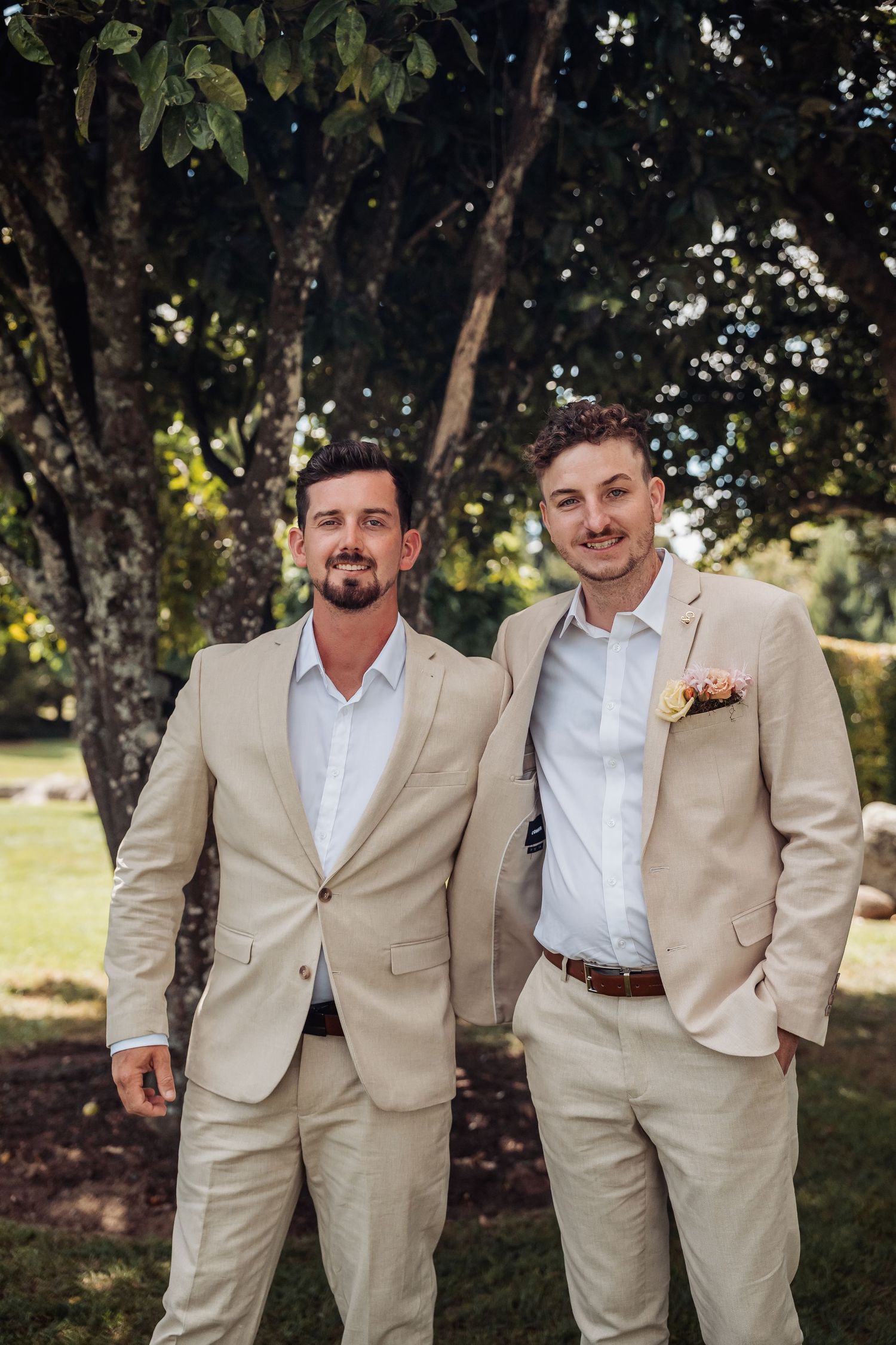 Two smiling groomsmen in tan suits posing outdoors with boutonniere on wedding day.