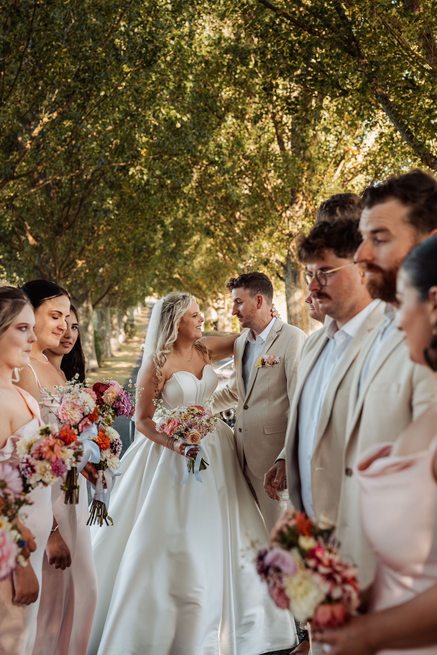 Wedding party gathers under lush green trees as bride and groom share a loving glance.