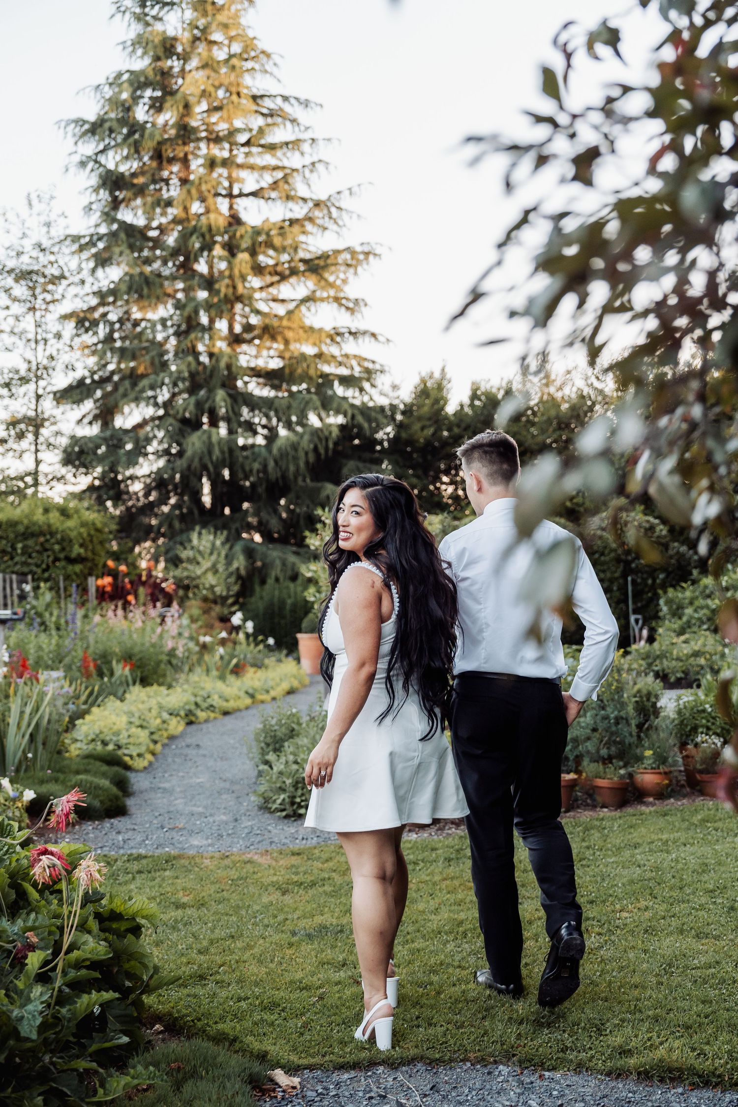A smiling bride in a white mini dress walks with her groom through a beautiful garden.