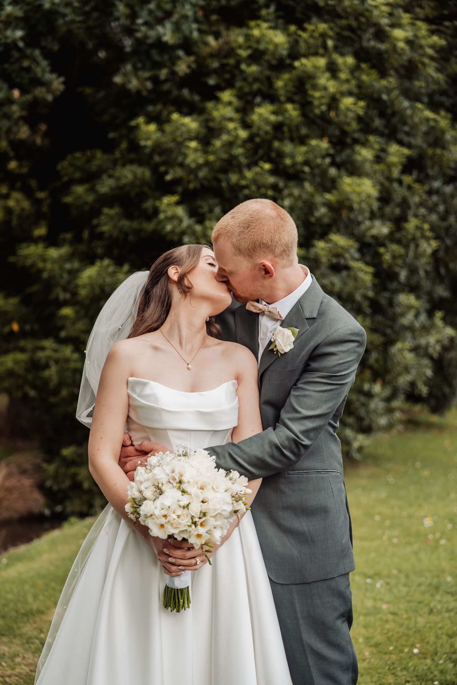 Groom kisses bride on cheek as she holds white floral bouquet in elegant strapless gown outdoors.