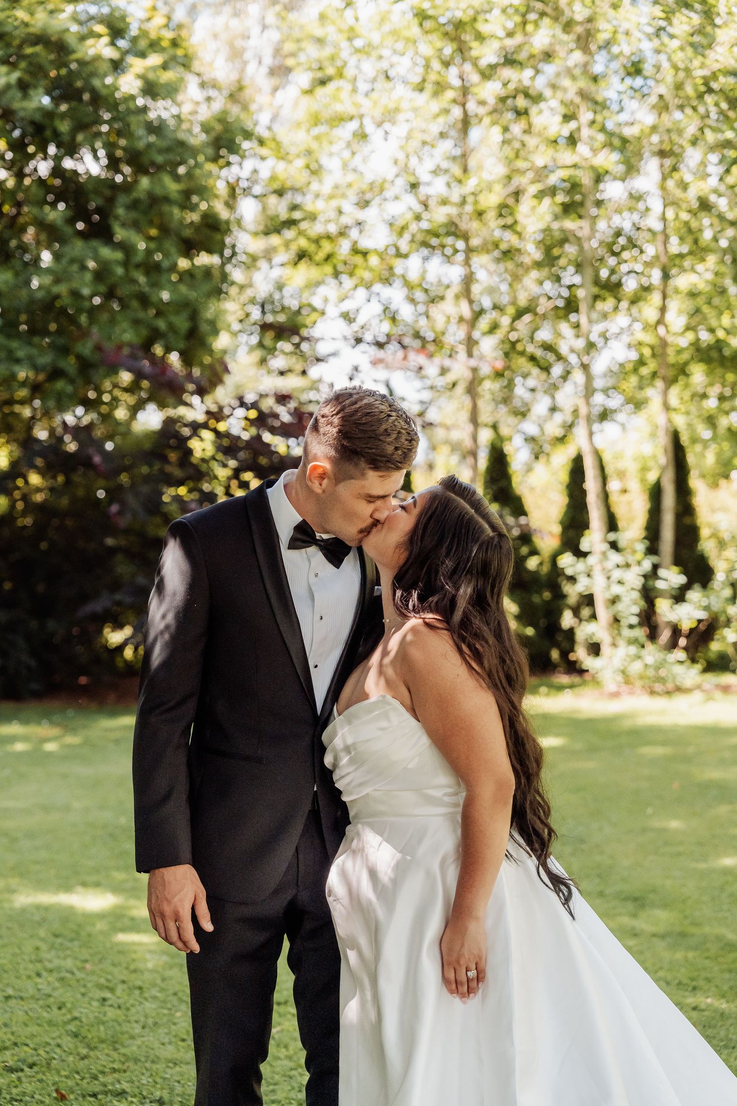 Bride and groom sharing a romantic kiss outdoors surrounded by lush green trees on their wedding day.
