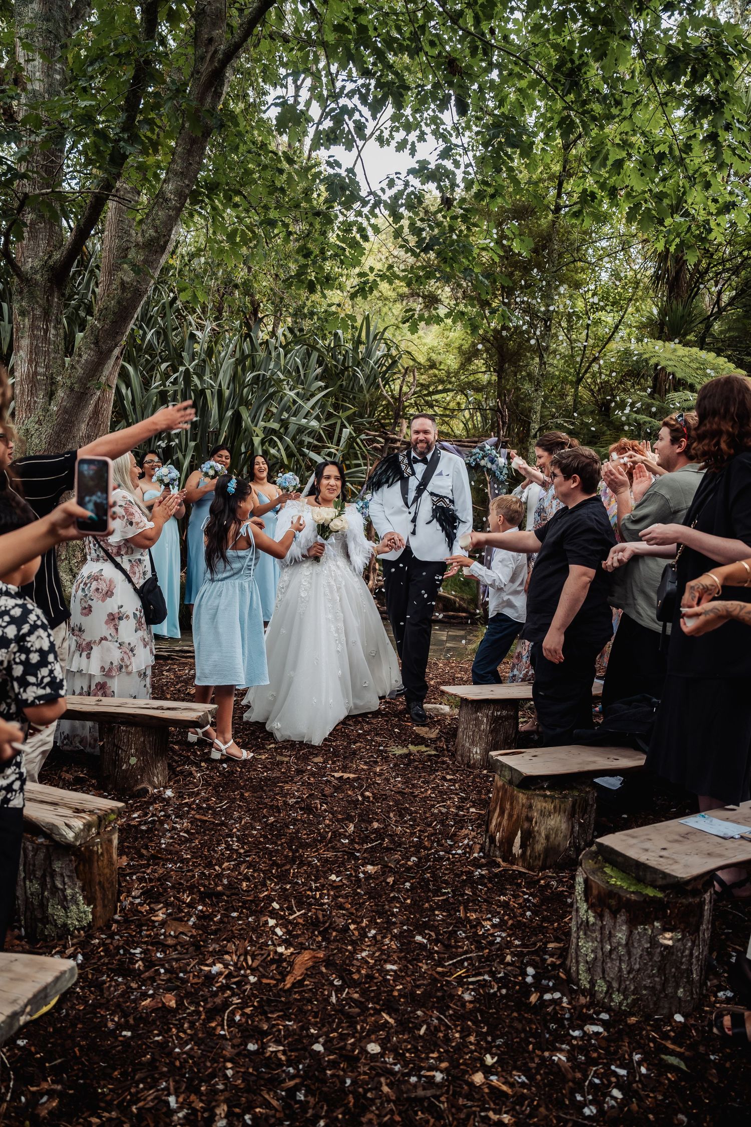 Joyful couple exits their outdoor forest wedding ceremony as guests celebrate along the wood chip aisle.