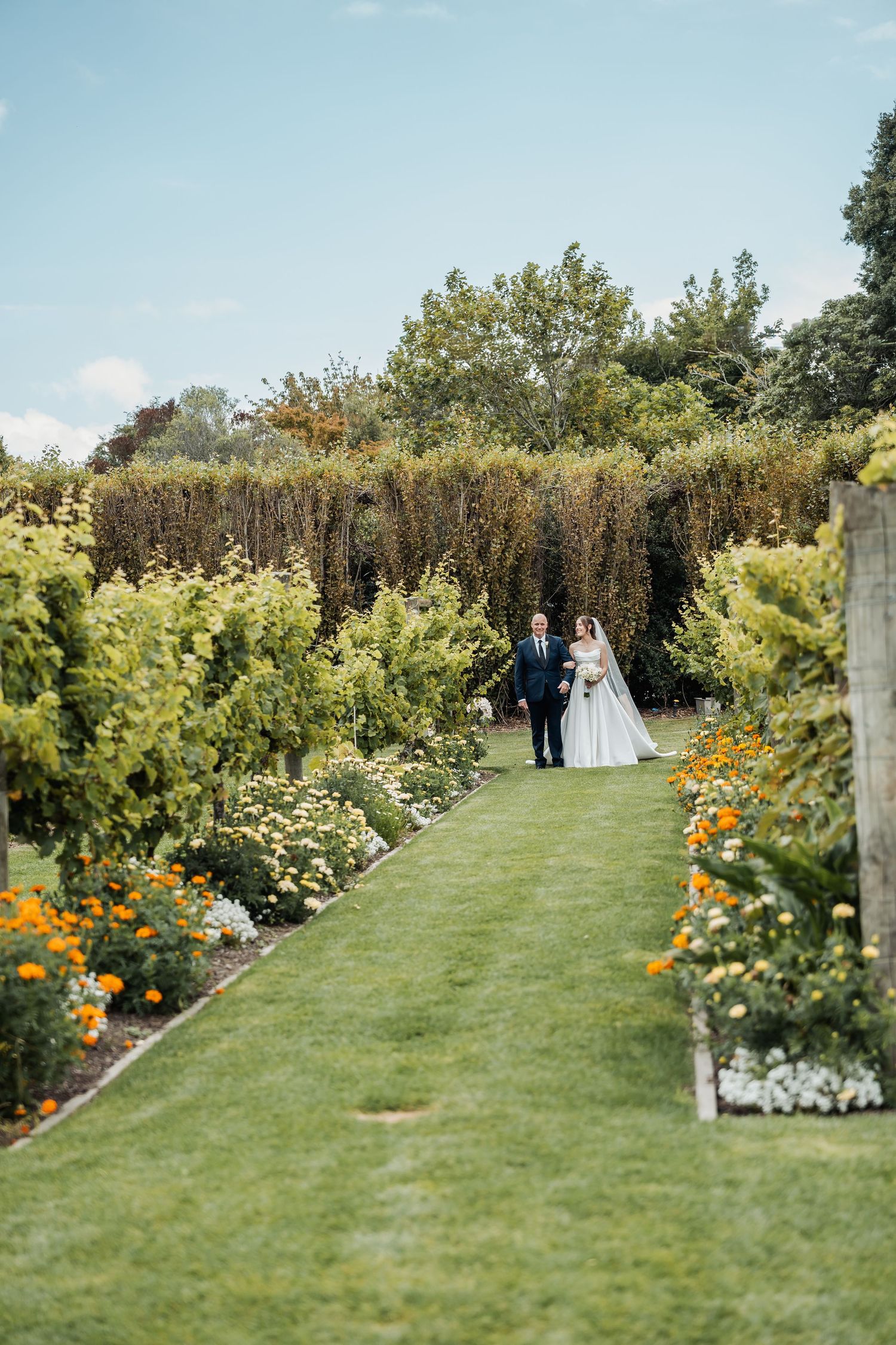 Bride and groom walk hand in hand along a lush garden path lined with colorful flowers on their wedding day.