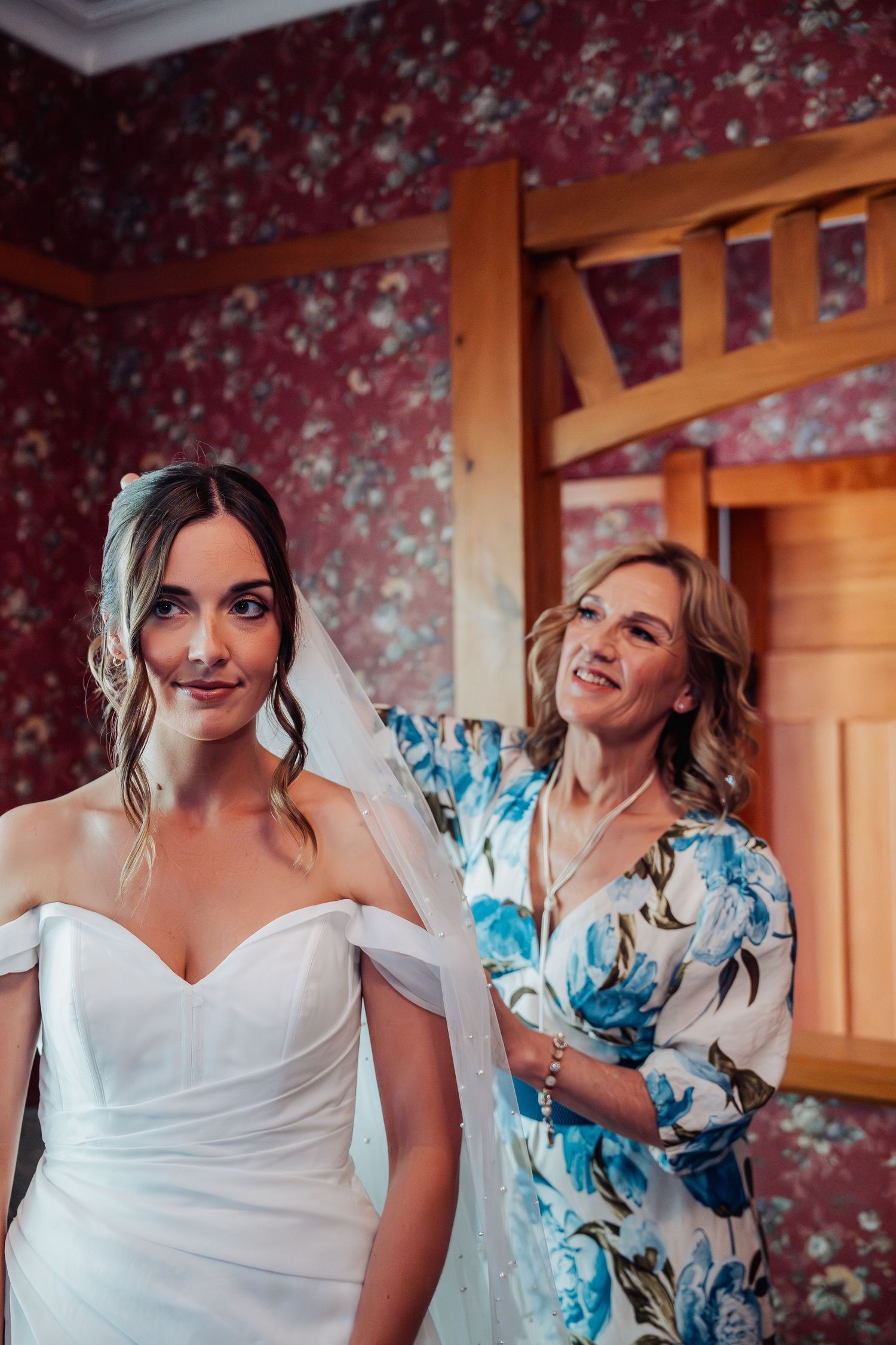 Bride in off-shoulder white gown smiles as woman in floral dress helps with veil.