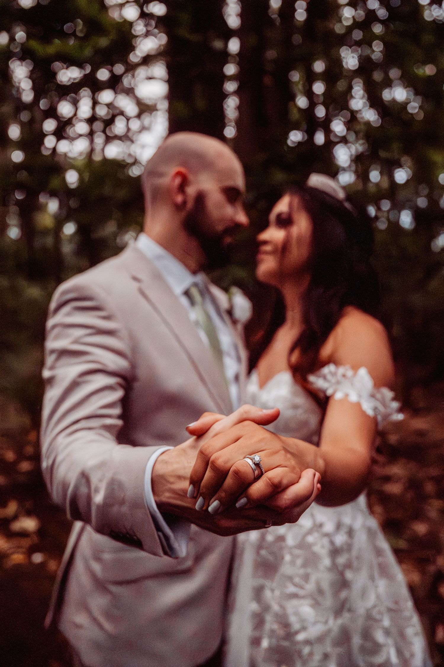 Newlyweds displaying wedding rings while holding hands in a sunlit forest setting.