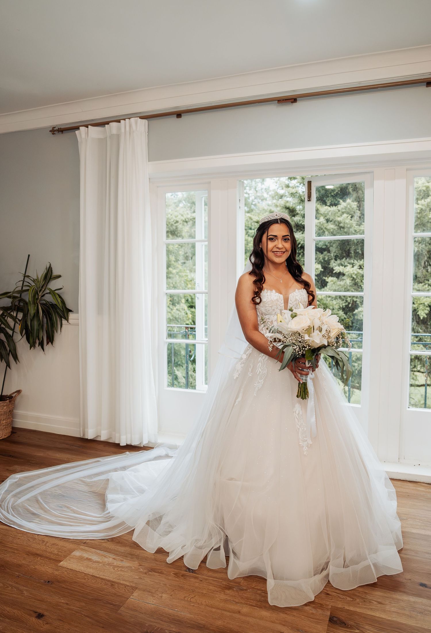 Bride in a strapless ballgown with long train holds white bouquet, standing before bright French windows.