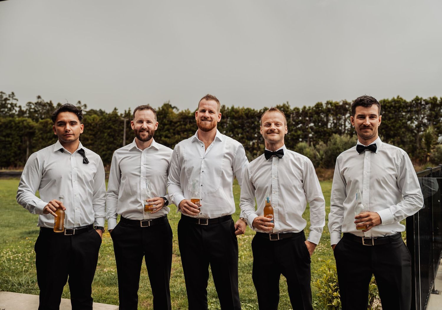Five groomsmen in white shirts and black bow ties smiling outdoors at a wedding celebration.