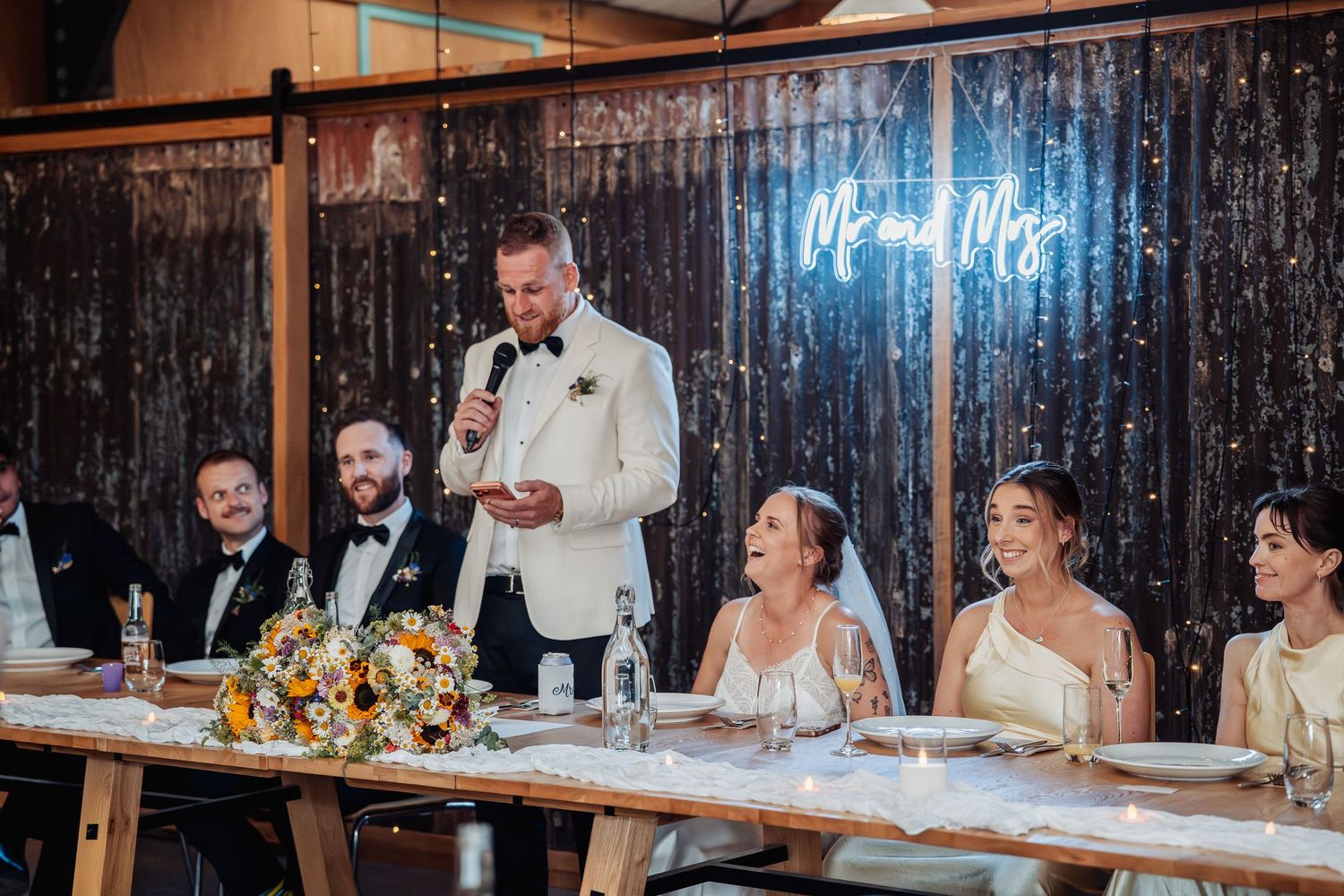 Groom in white jacket giving speech at wedding reception with neon Mr and Mrs sign.
