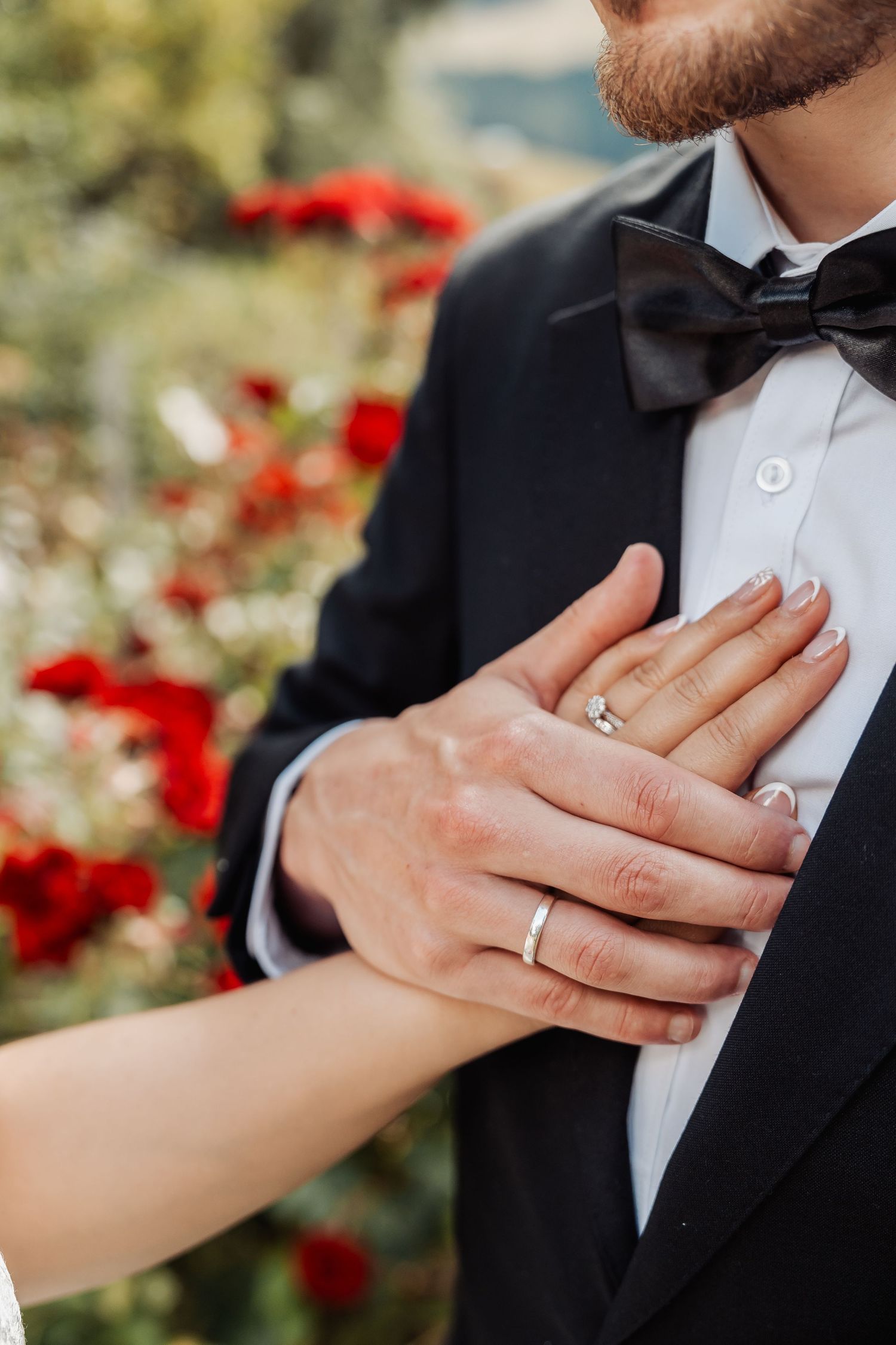 Close-up of bride and groom hands showing wedding rings, groom wearing black tuxedo with bow tie.