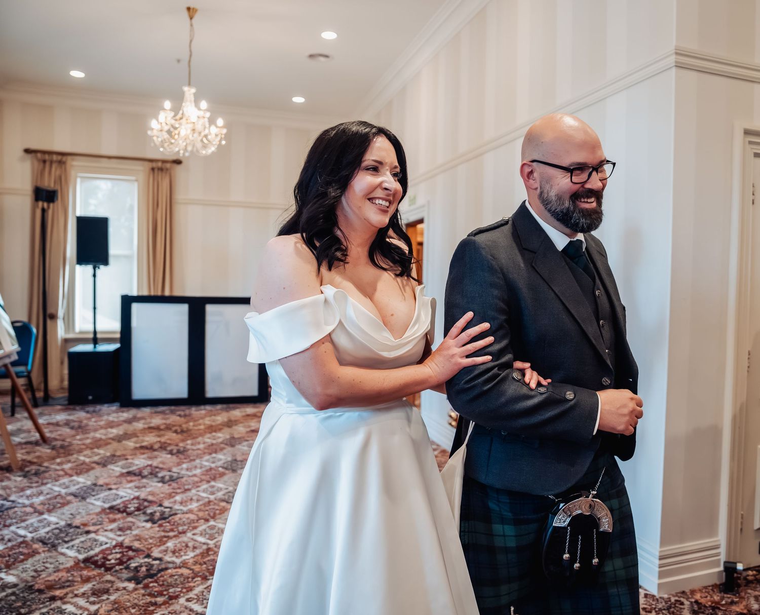 Bride in white off-shoulder gown and groom in Scottish kilt smiling during wedding reception.