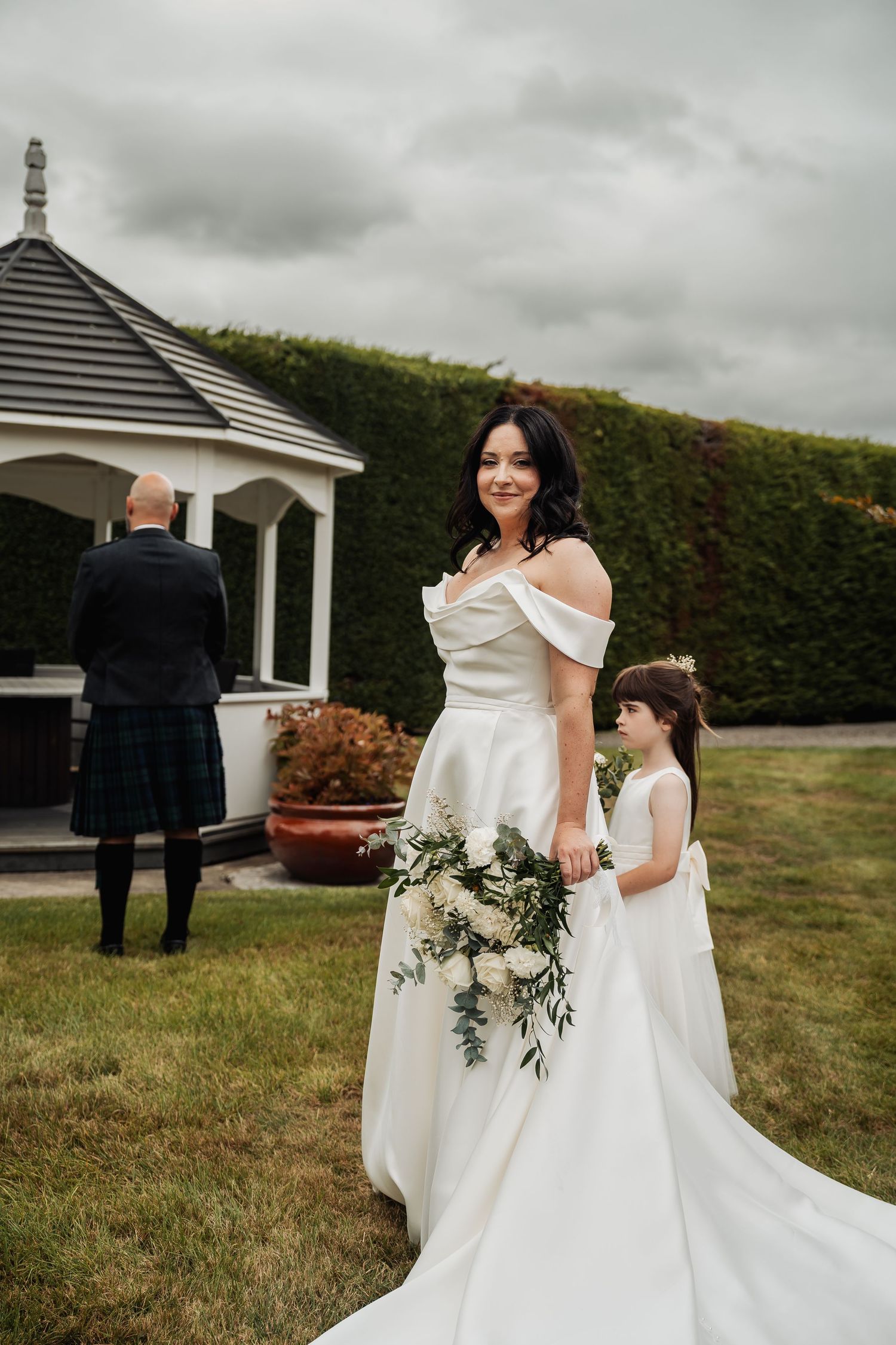 Smiling bride in elegant white off-shoulder gown holds bouquet with flower girl outdoors near a gazebo.