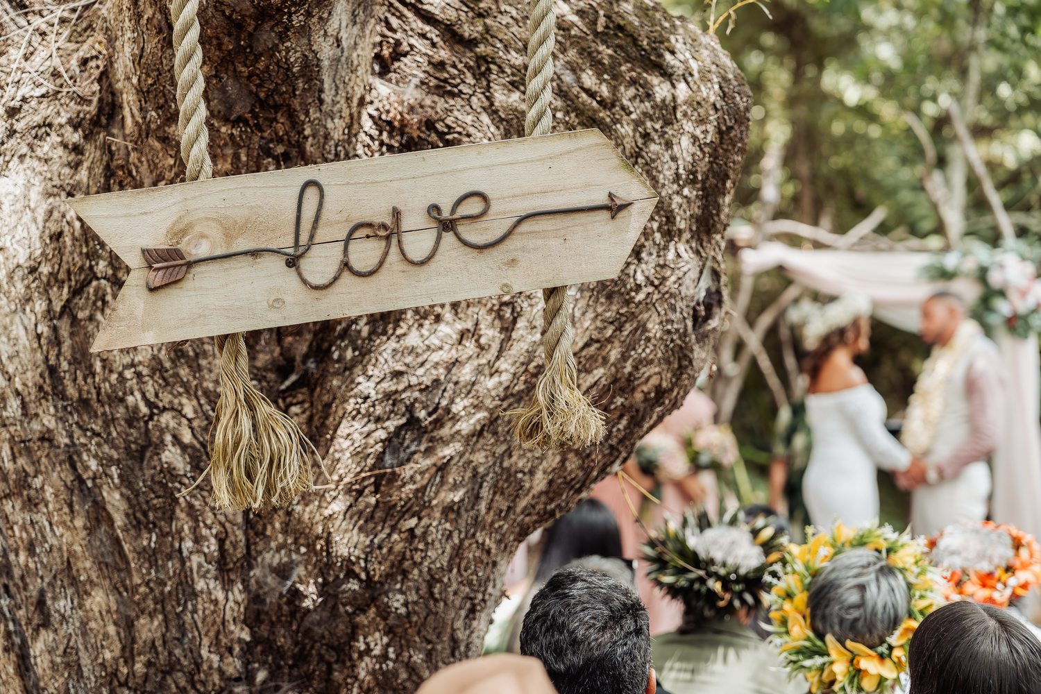 Rustic wooden 'love' sign hanging from rope with wedding celebration in background.