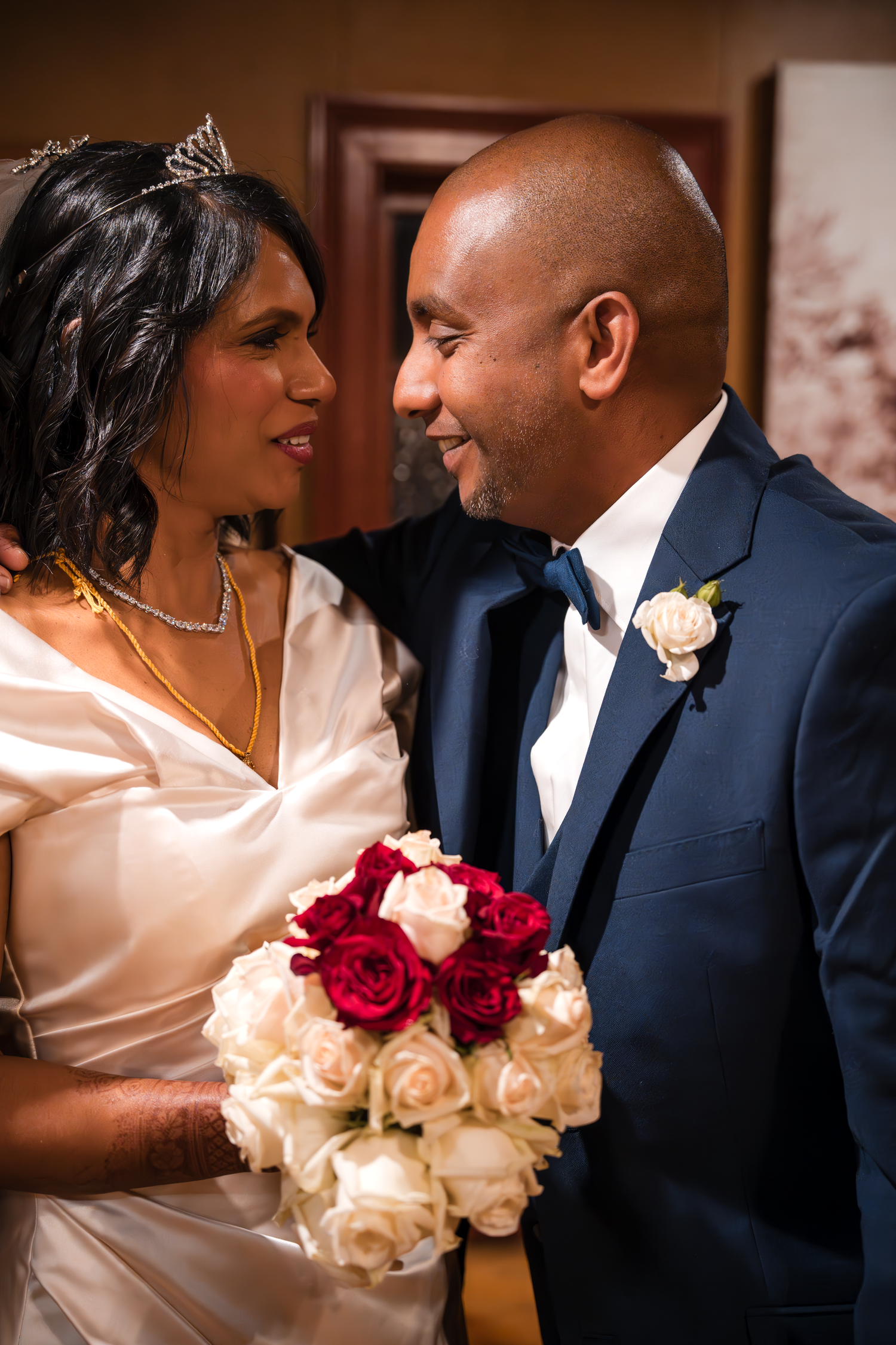 A bride in a white dress and tiara holds a red and white rose bouquet next to her groom in a navy suit.