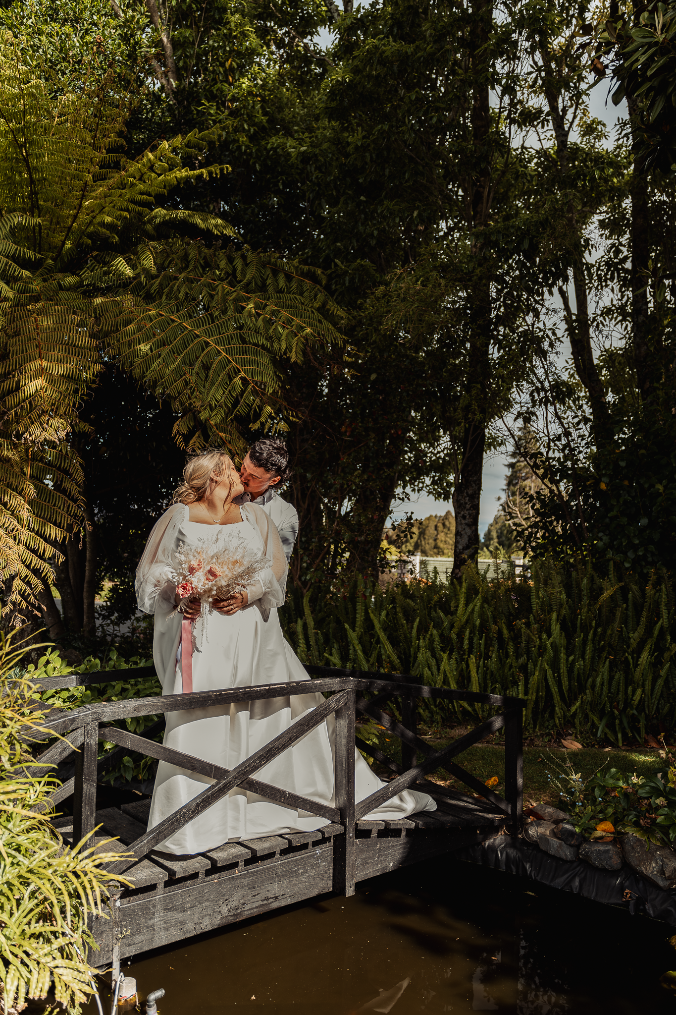 Newlyweds embrace on rustic bridge surrounded by ferns and garden landscaping.