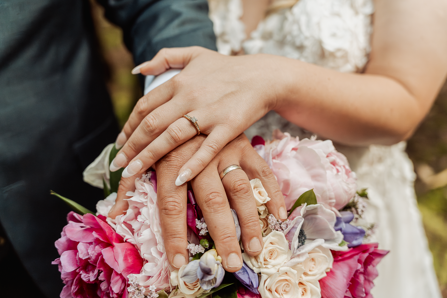Close-up of wedding rings and hands resting on a pink and white floral bouquet.