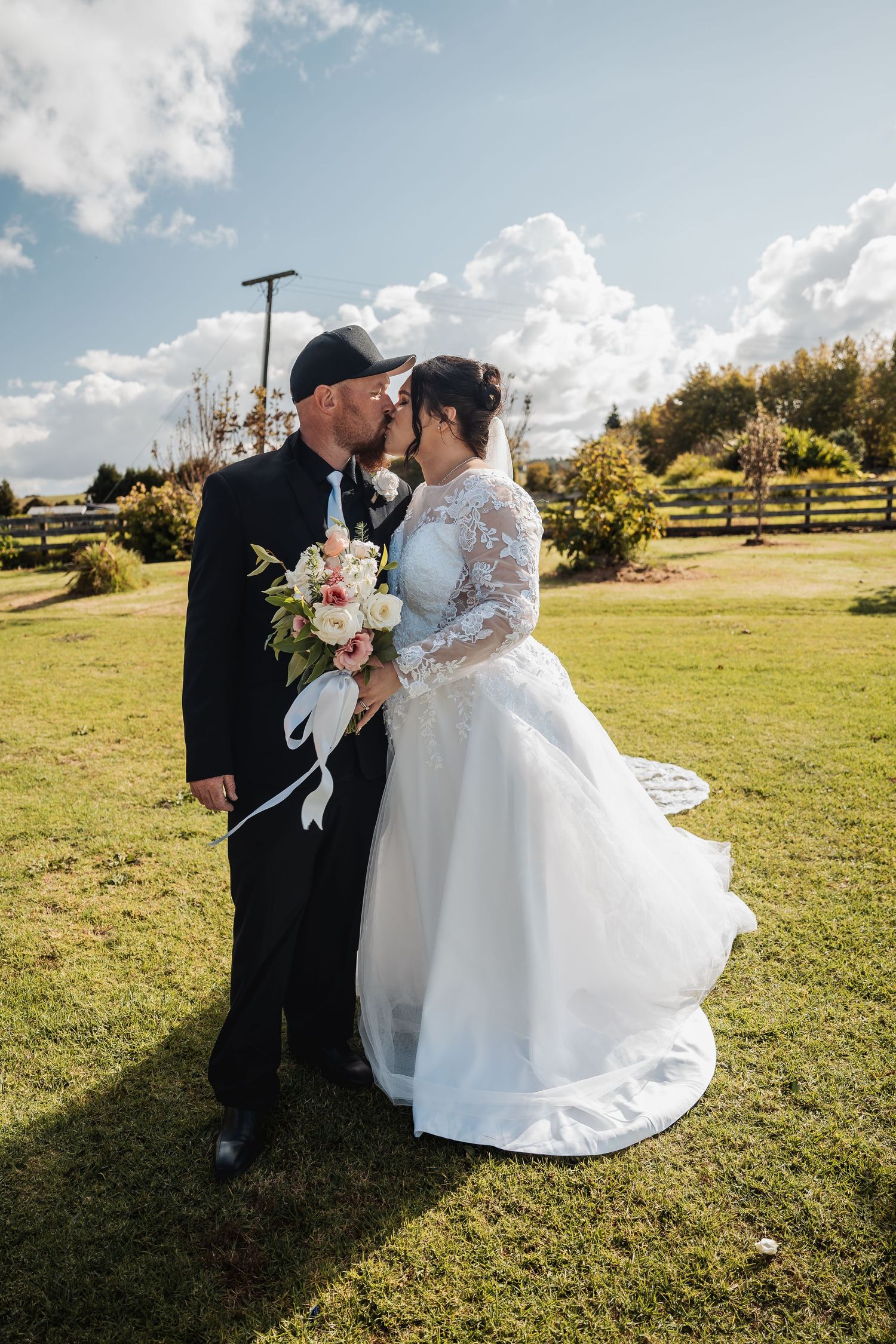 A newlywed couple shares a romantic kiss while holding a bouquet in a sunny garden setting.