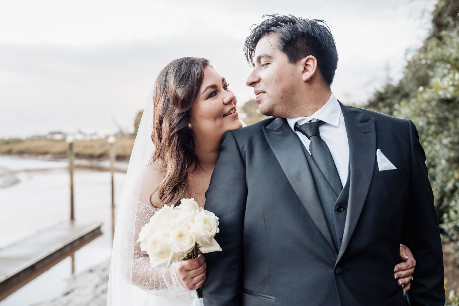 A happy newlywed couple shares an intimate moment on a waterfront dock while the bride holds a white bouquet.