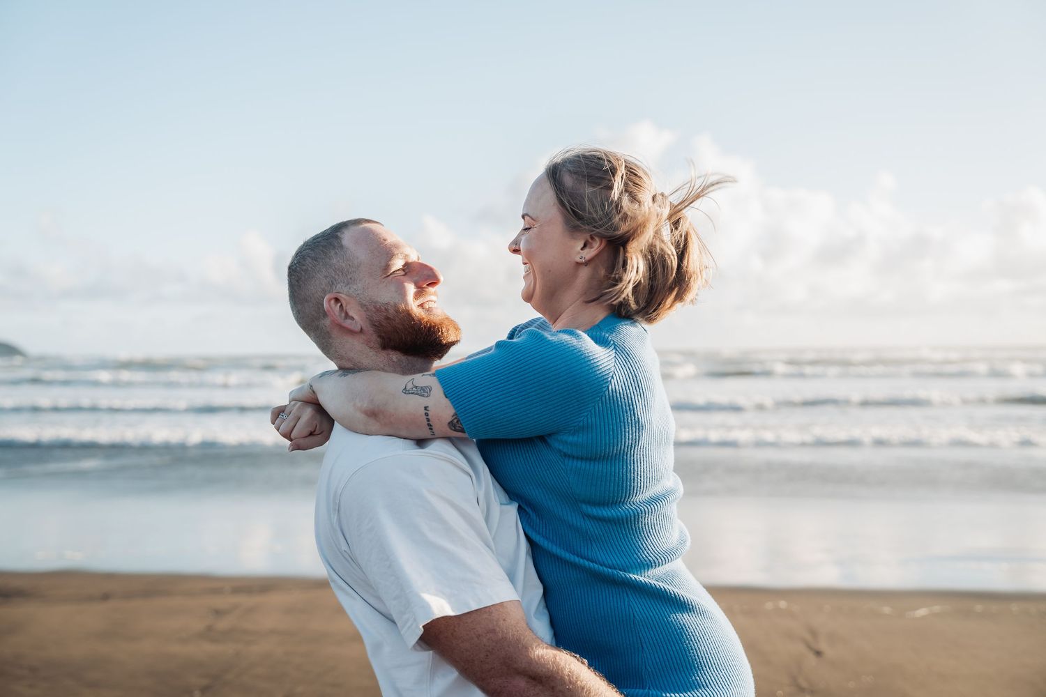 A joyful couple embracing and laughing together on a beach with the ocean and cloudy sky behind them.