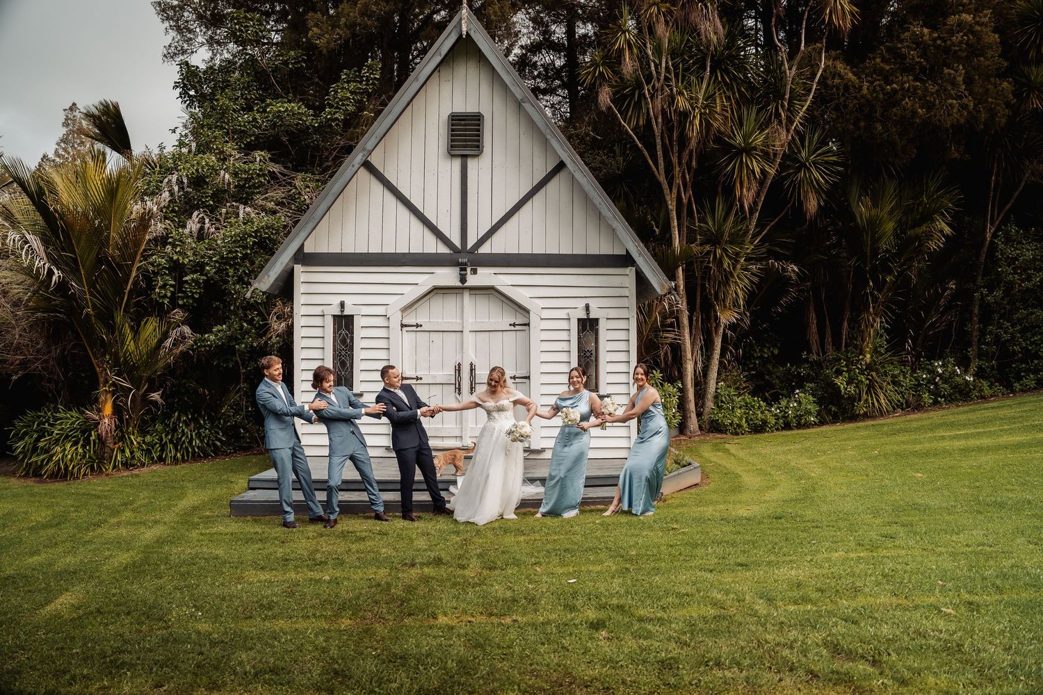 Wedding party poses in front of charming white chapel on grassy lawn.