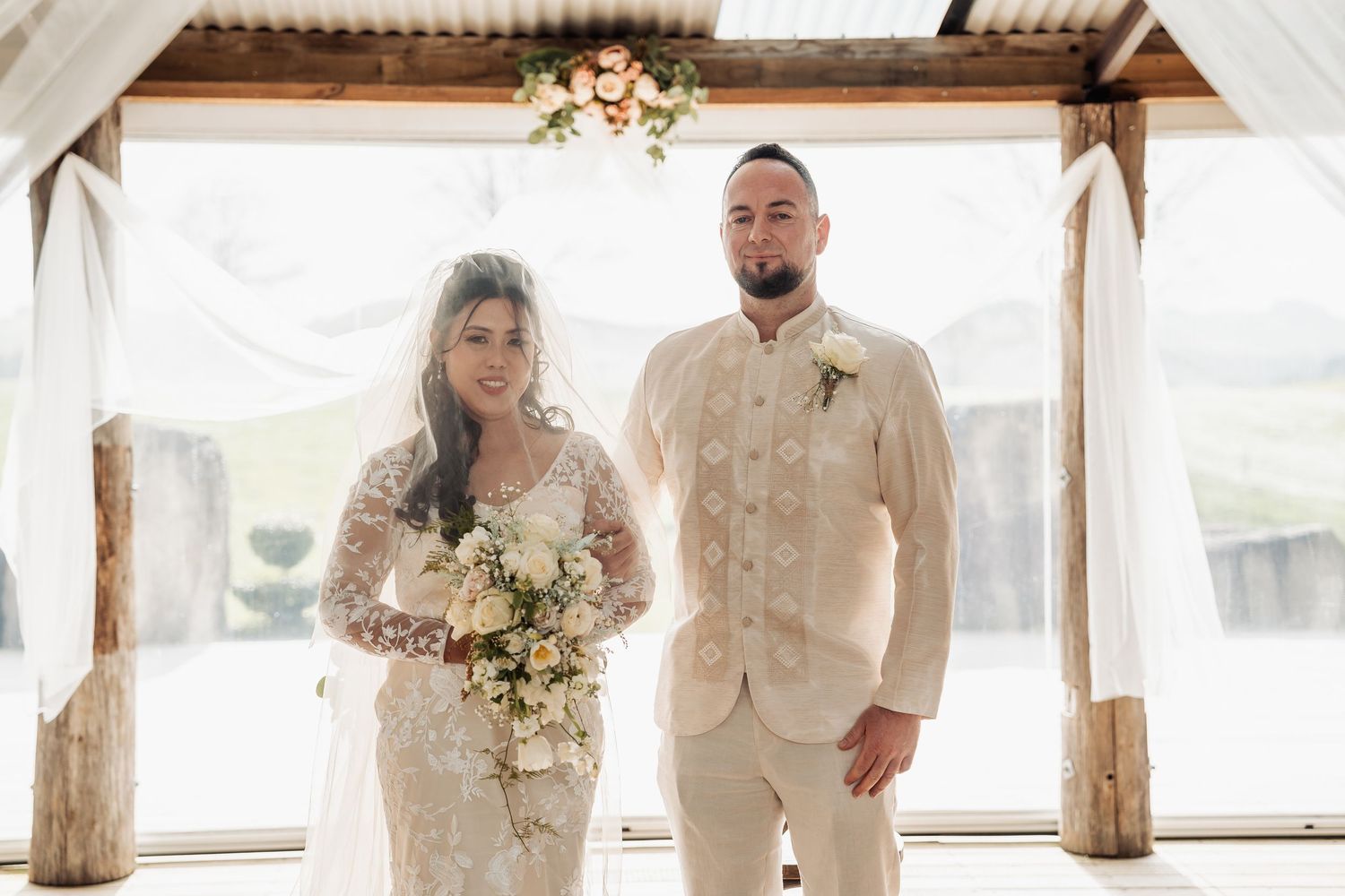 A couple stands under a rustic wooden pavilion decorated with floral arrangements on their wedding day.