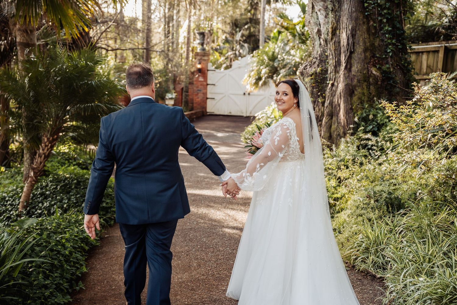 A bride in white and groom in navy walk hand in hand down a garden path.