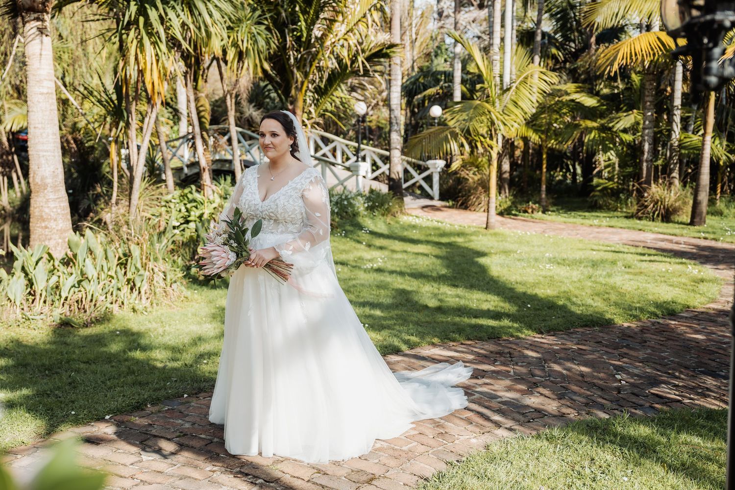 A couple in wedding attire walks hand in hand through a lush tropical garden path lined with palm trees.