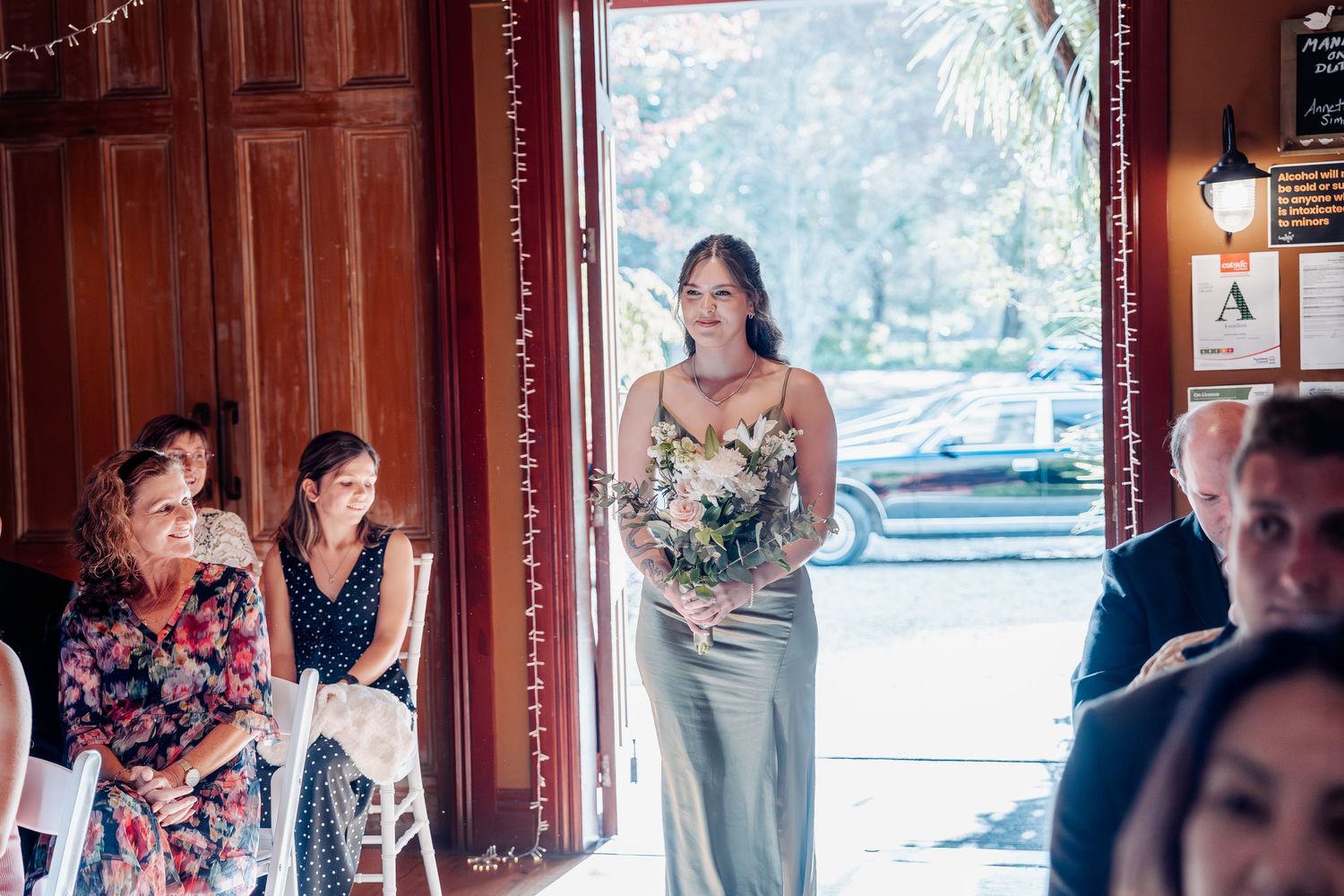 A bridesmaid in a silk sage green dress holds a bouquet while standing in a rustic wooden doorway during a wedding.