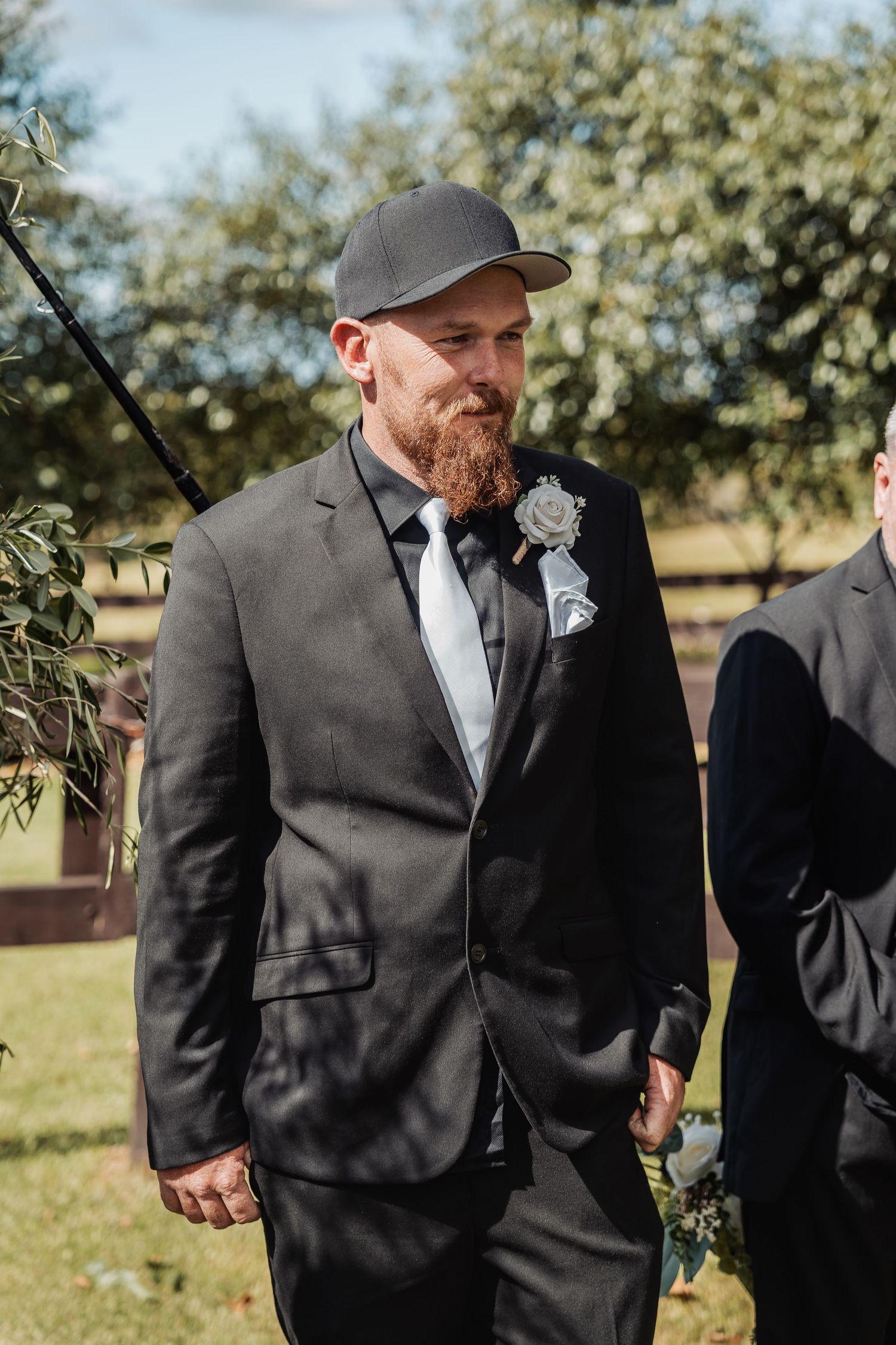 Man in grey suit and baseball cap stands outdoors at formal event with boutonniere.