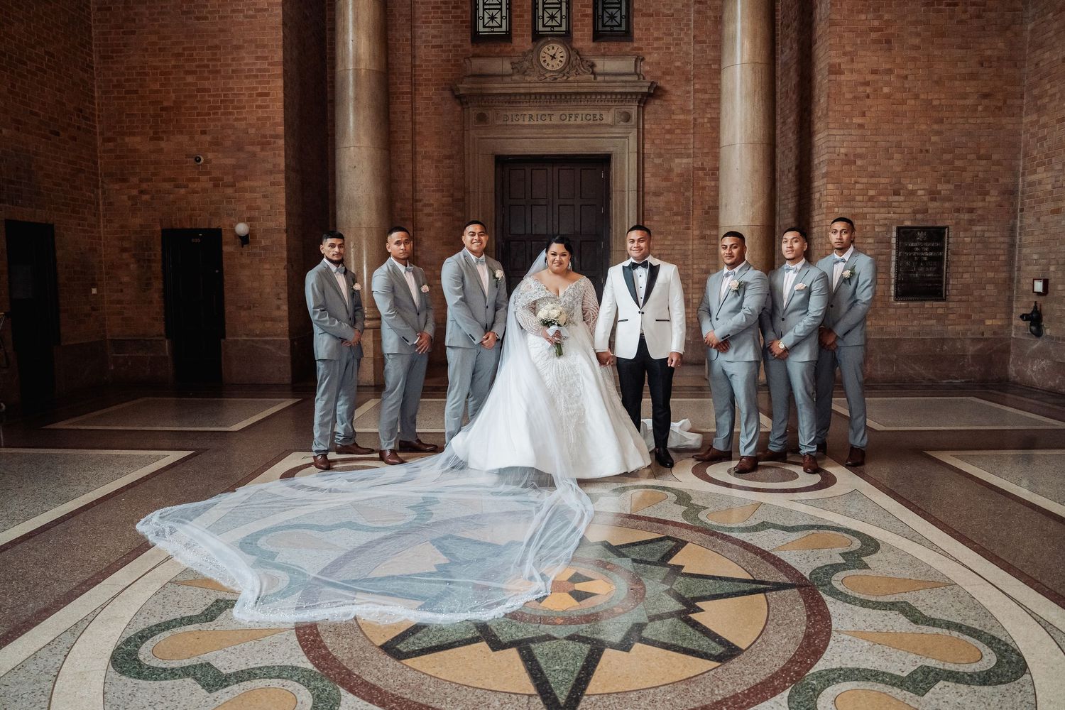 Wedding party poses on ornate floor mosaic pattern in historic brick building interior.