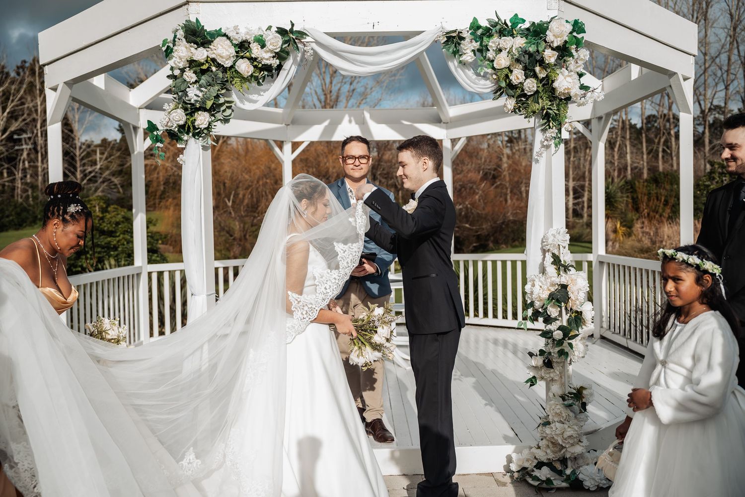 Outdoor wedding ceremony scene with white gazebo decorated in greenery and floral garlands.
