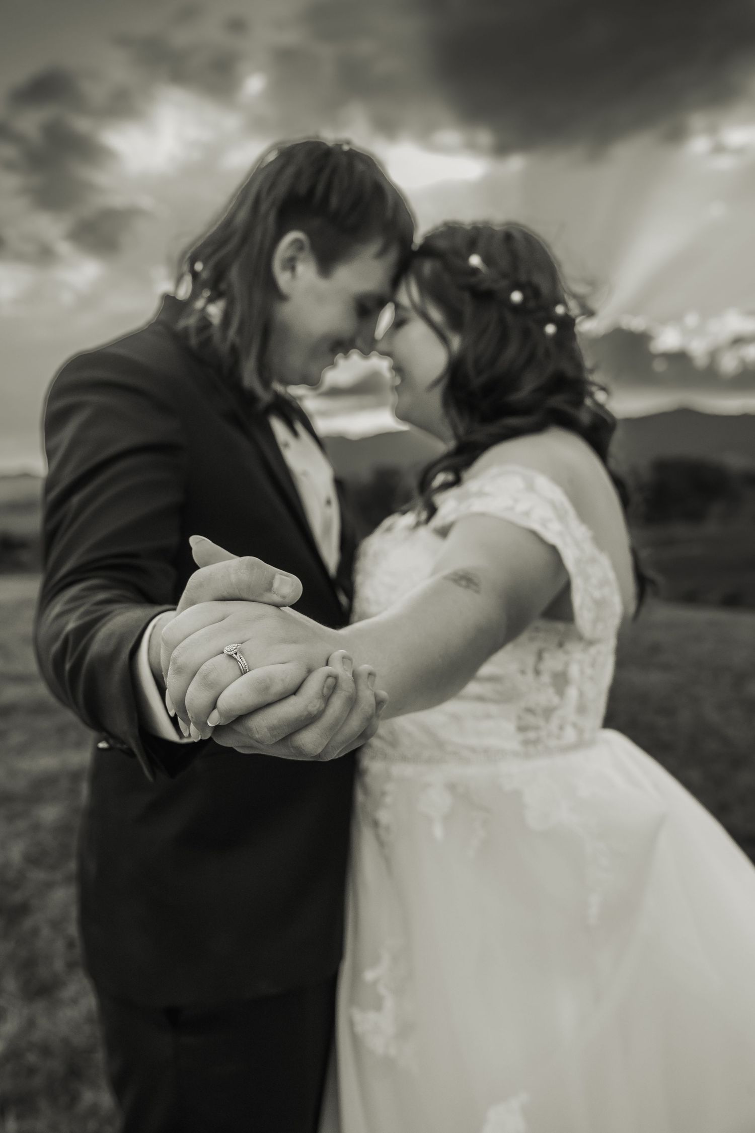 Black and white photo of bride and groom holding hands and touching foreheads under dramatic sky.