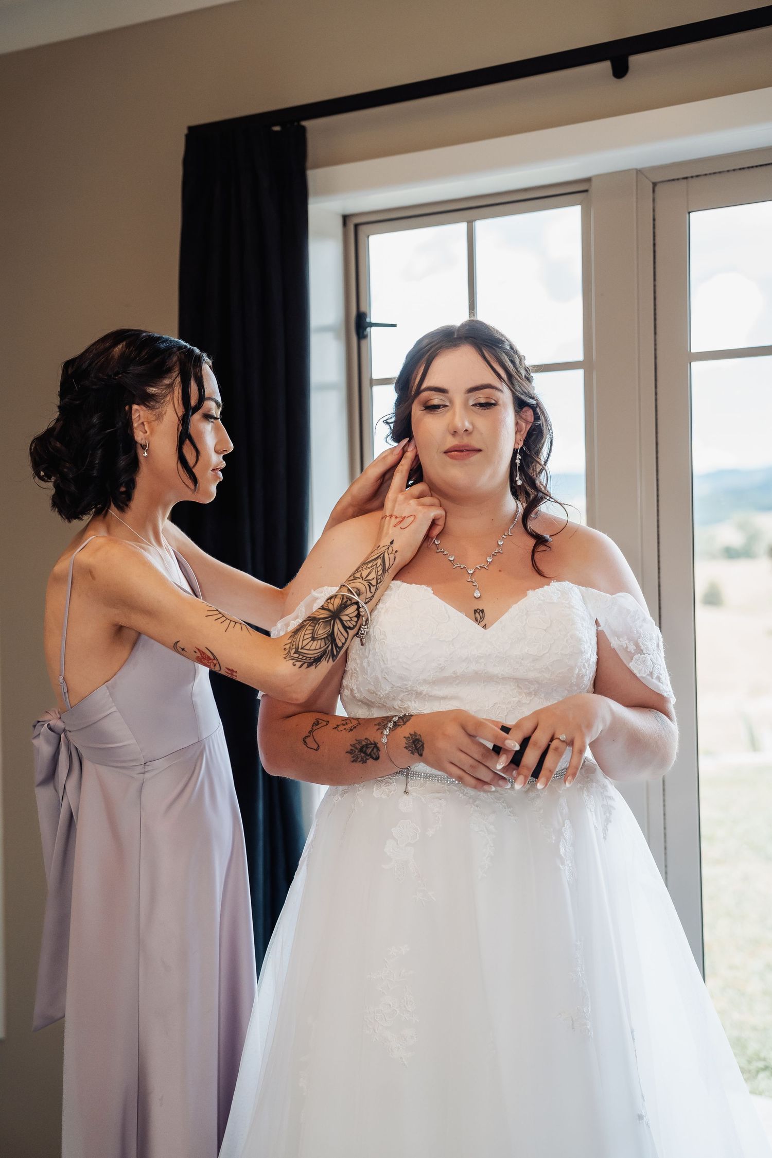 Bridesmaid with henna tattoos helping bride in white off-shoulder gown with necklace near bright window.
