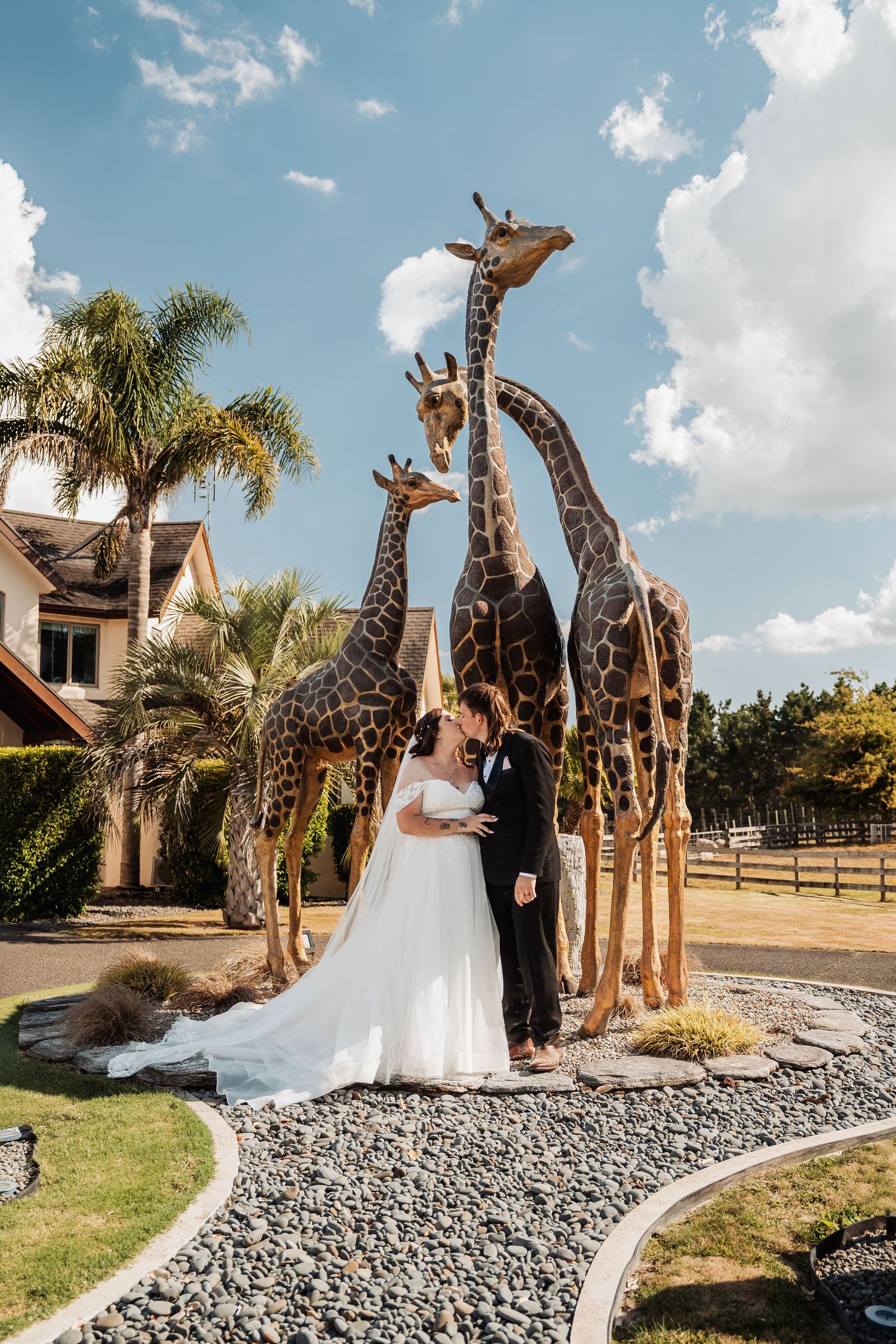 Bride and groom share a kiss surrounded by tall giraffe sculptures in a scenic outdoor garden setting.