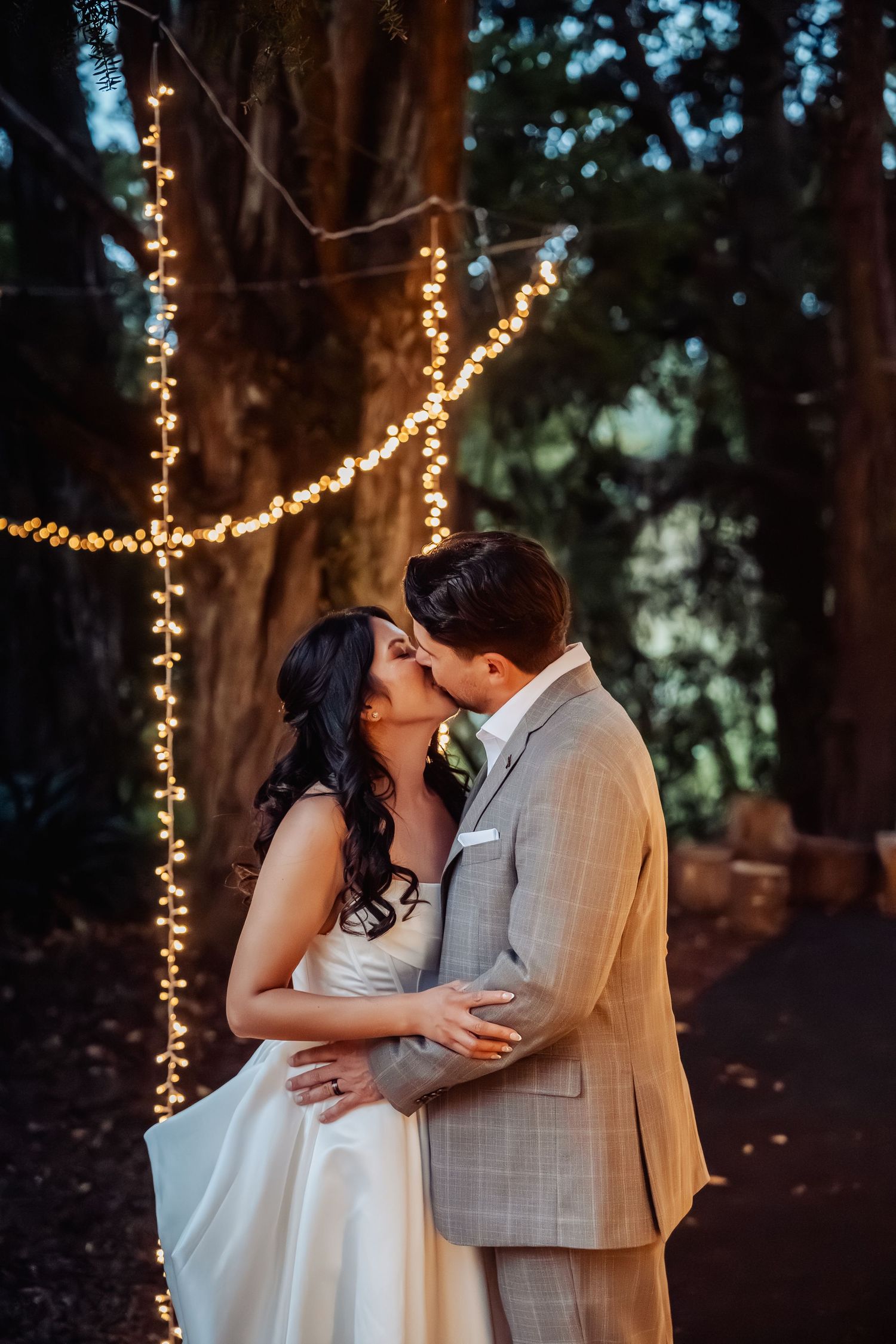Bride and groom share romantic kiss under twinkling fairy lights in wooded outdoor wedding setting.