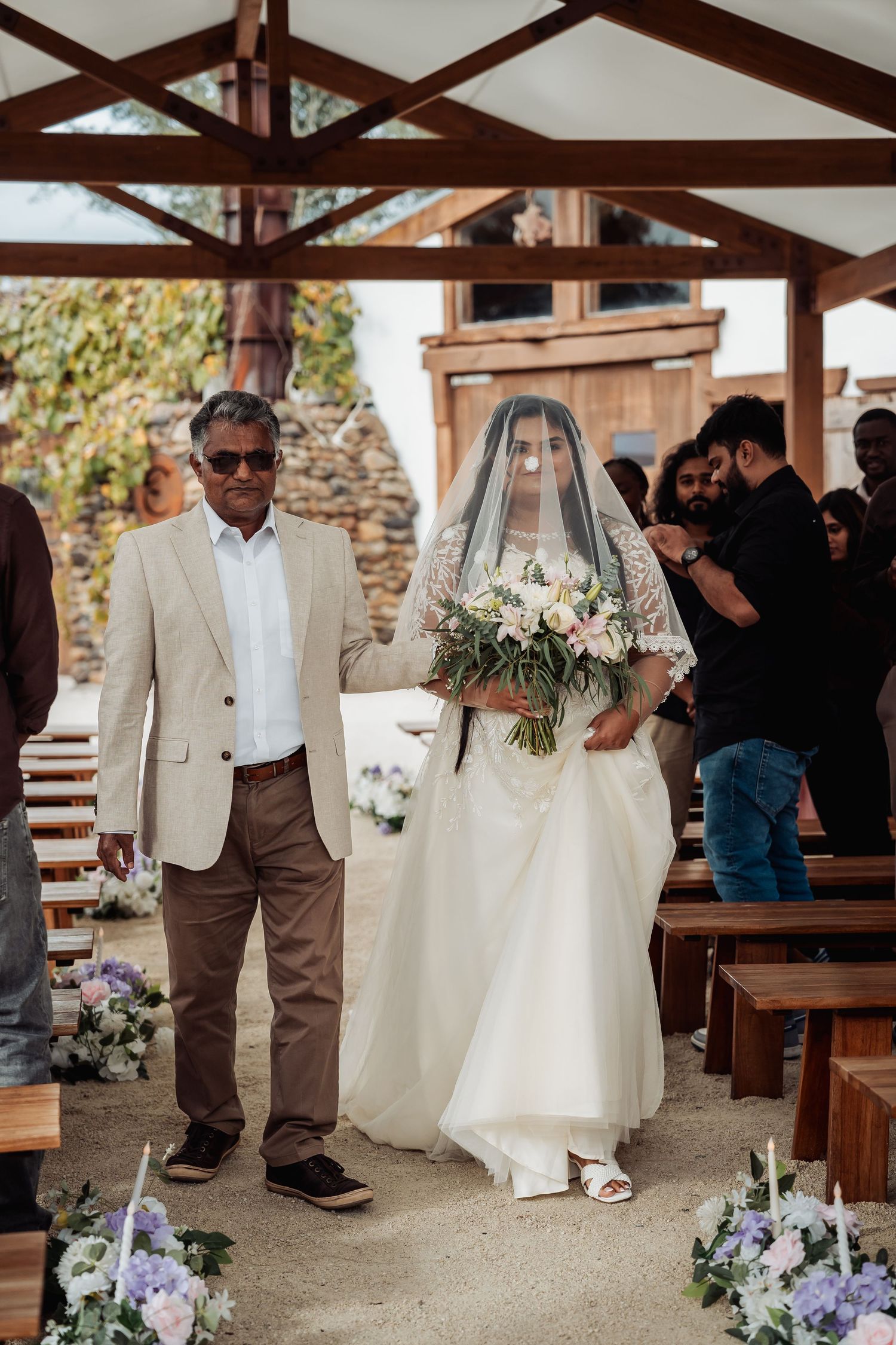 Bride in white gown and veil walks down aisle with father at outdoor wedding ceremony.