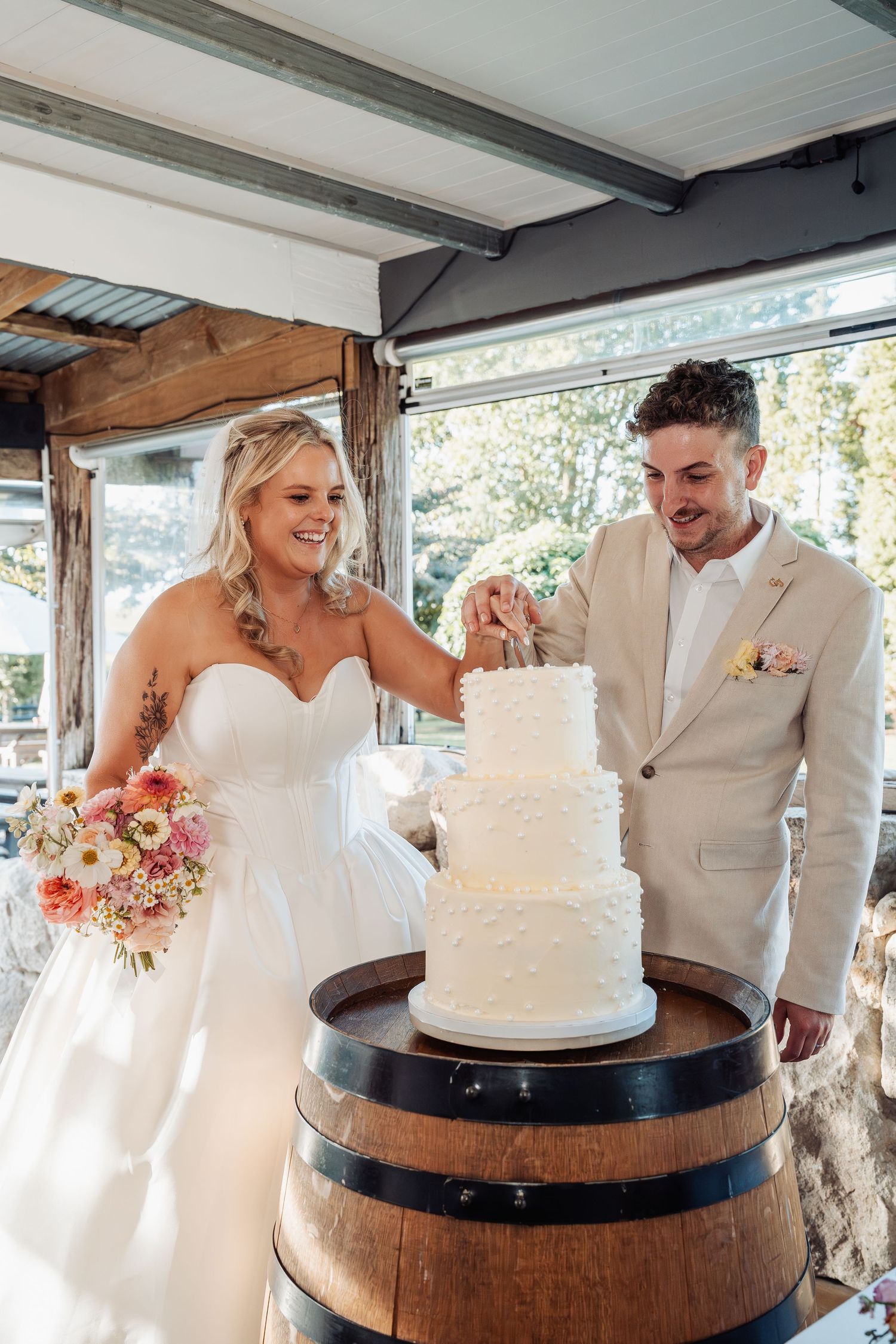 Bride and groom cut a three-tier white wedding cake displayed on a rustic wooden barrel.