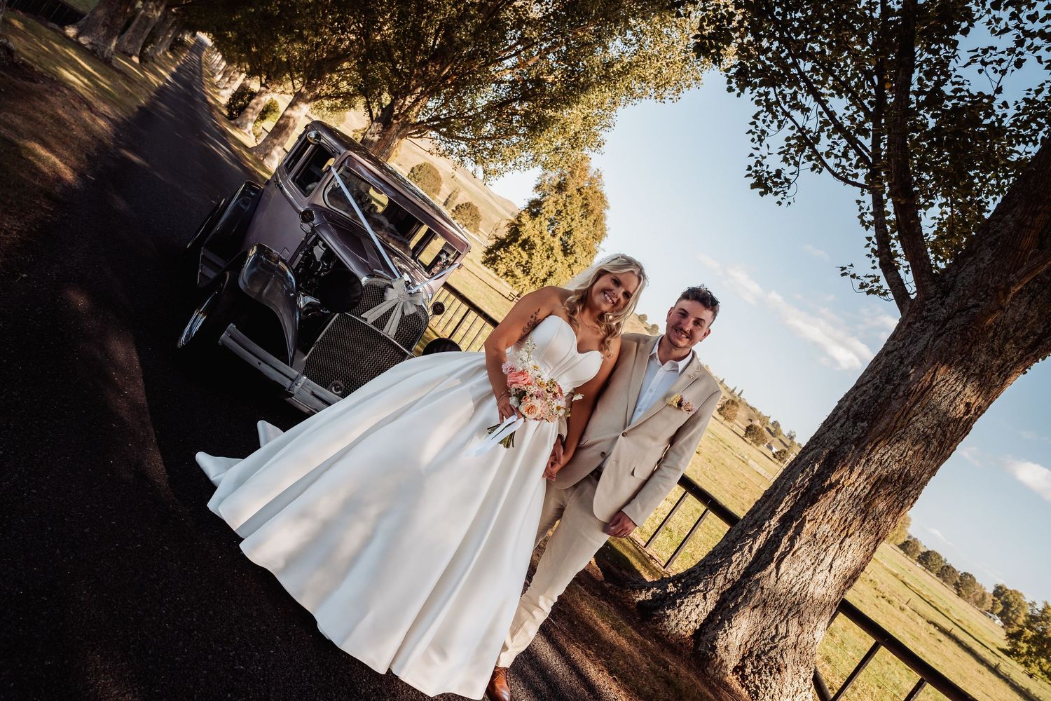 Bride in white gown and groom in beige suit pose together near a vintage car under trees at golden hour.