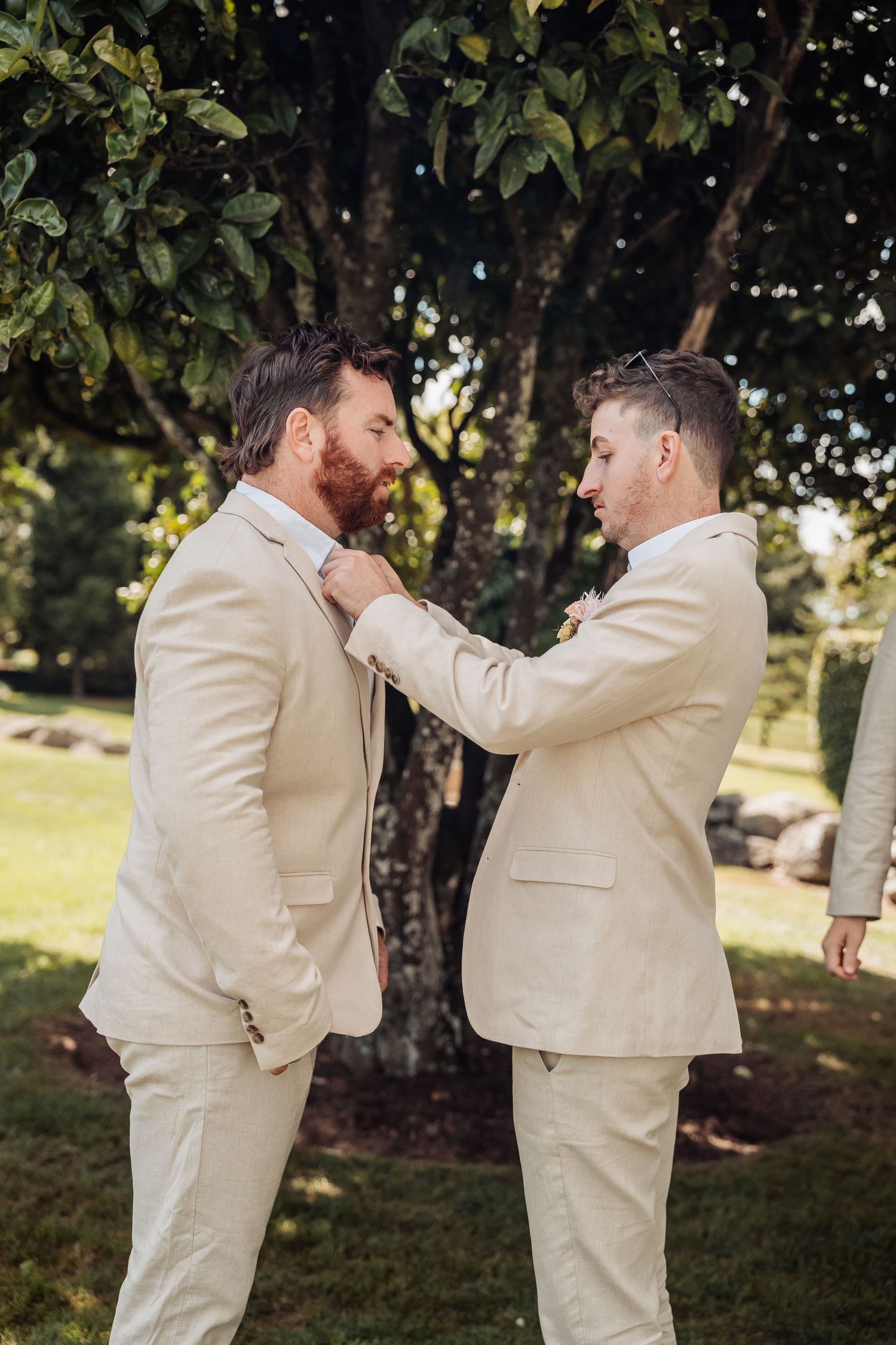 Groomsman adjusting groom's tie outdoors in matching beige suits before wedding ceremony.