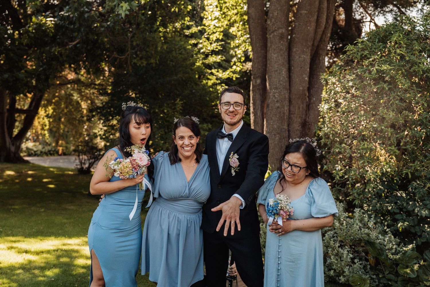 Groom in a black tuxedo posing with three bridesmaids in light blue dresses in a lush garden setting.