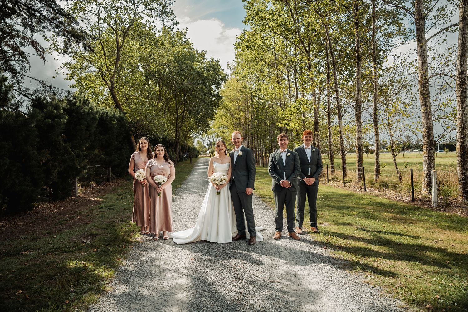 Wedding party posing on a gravel path surrounded by tall trees, featuring bride in white gown and bridesmaids in dusty rose dresses.