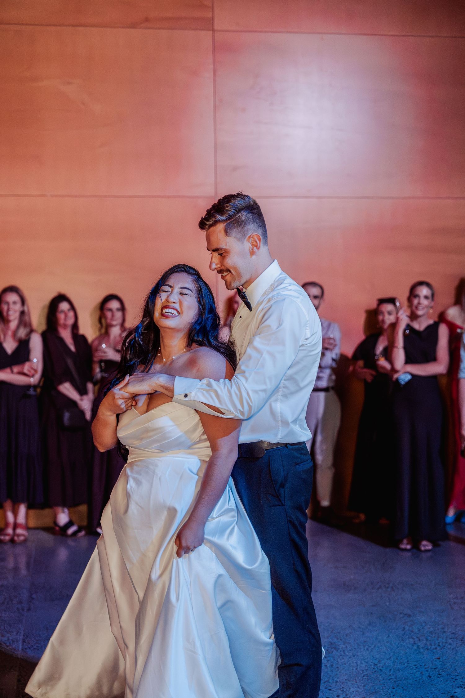 Laughing bride and groom share their first dance on the reception floor surrounded by guests in black attire.