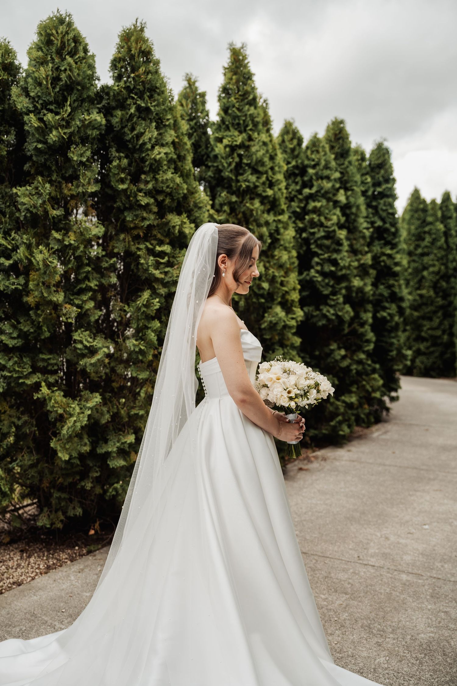 Beautiful bride in flowing white wedding dress and long veil poses with bouquet beside lush green trees.