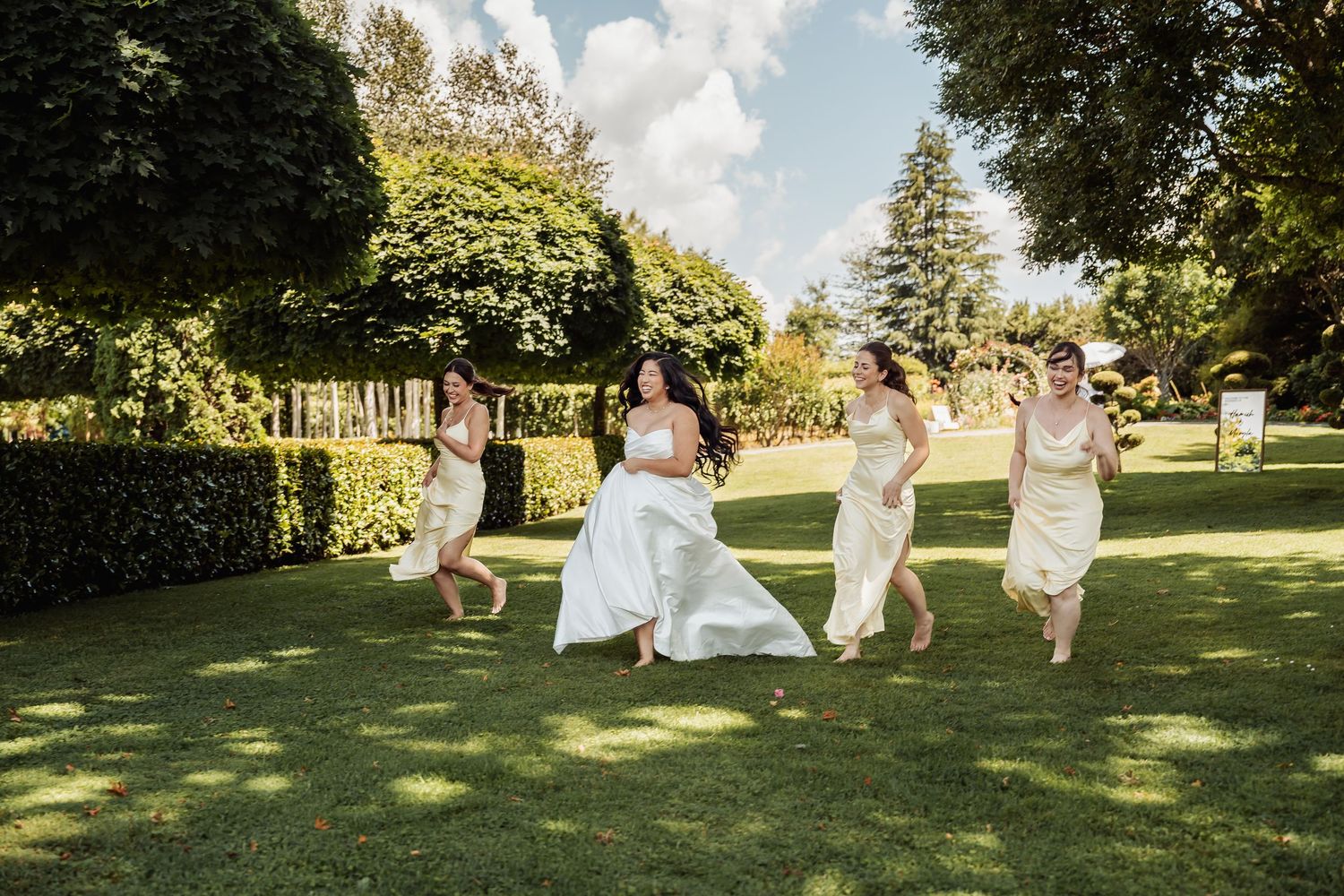Bride and bridesmaids in white dresses running joyfully across lush green garden lawn on sunny wedding day.