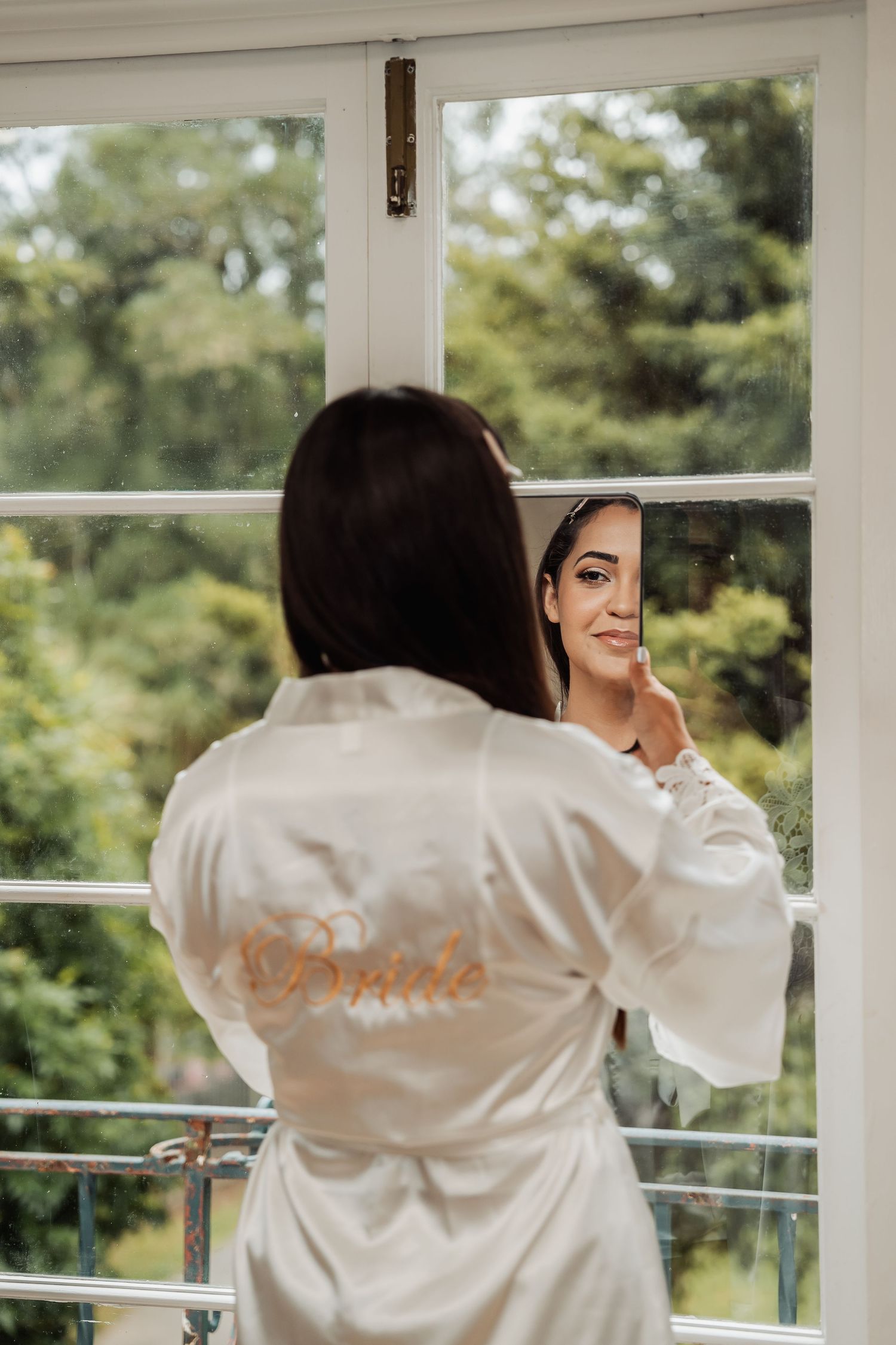 Bride in white satin Bride robe admiring her reflection in window mirror before wedding ceremony.