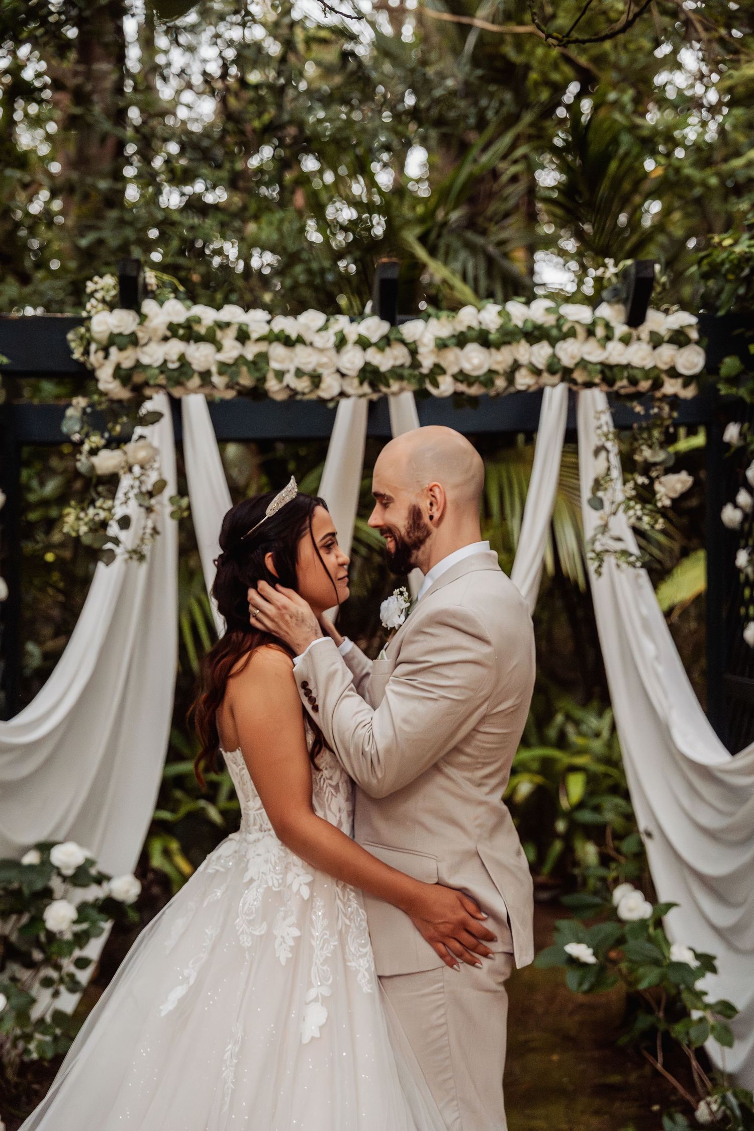 Bride and groom share intimate moment under white floral arch decorated with roses in garden ceremony.
